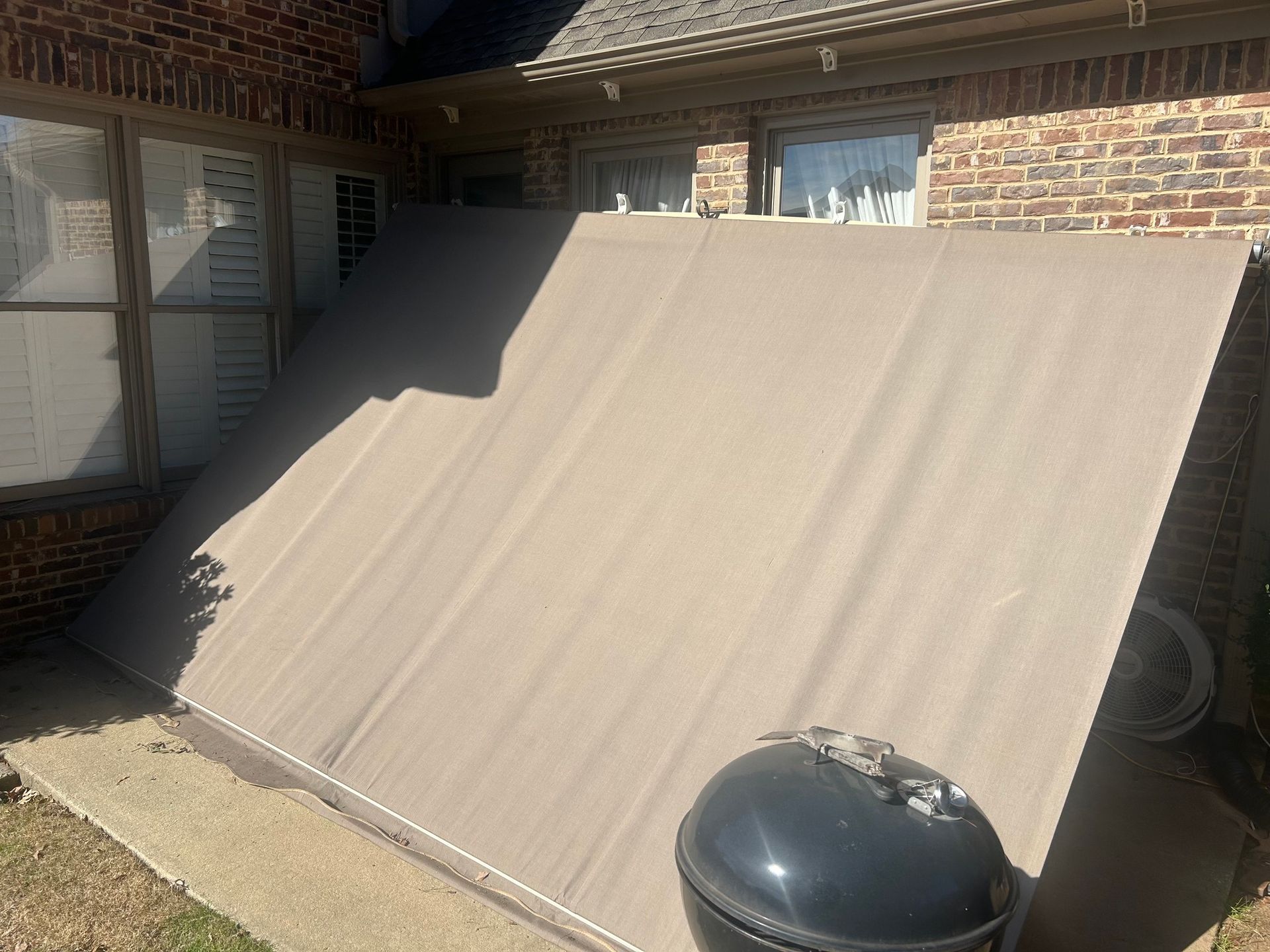 Beige retractable awning leaned against brick wall. Charcoal grill in foreground.