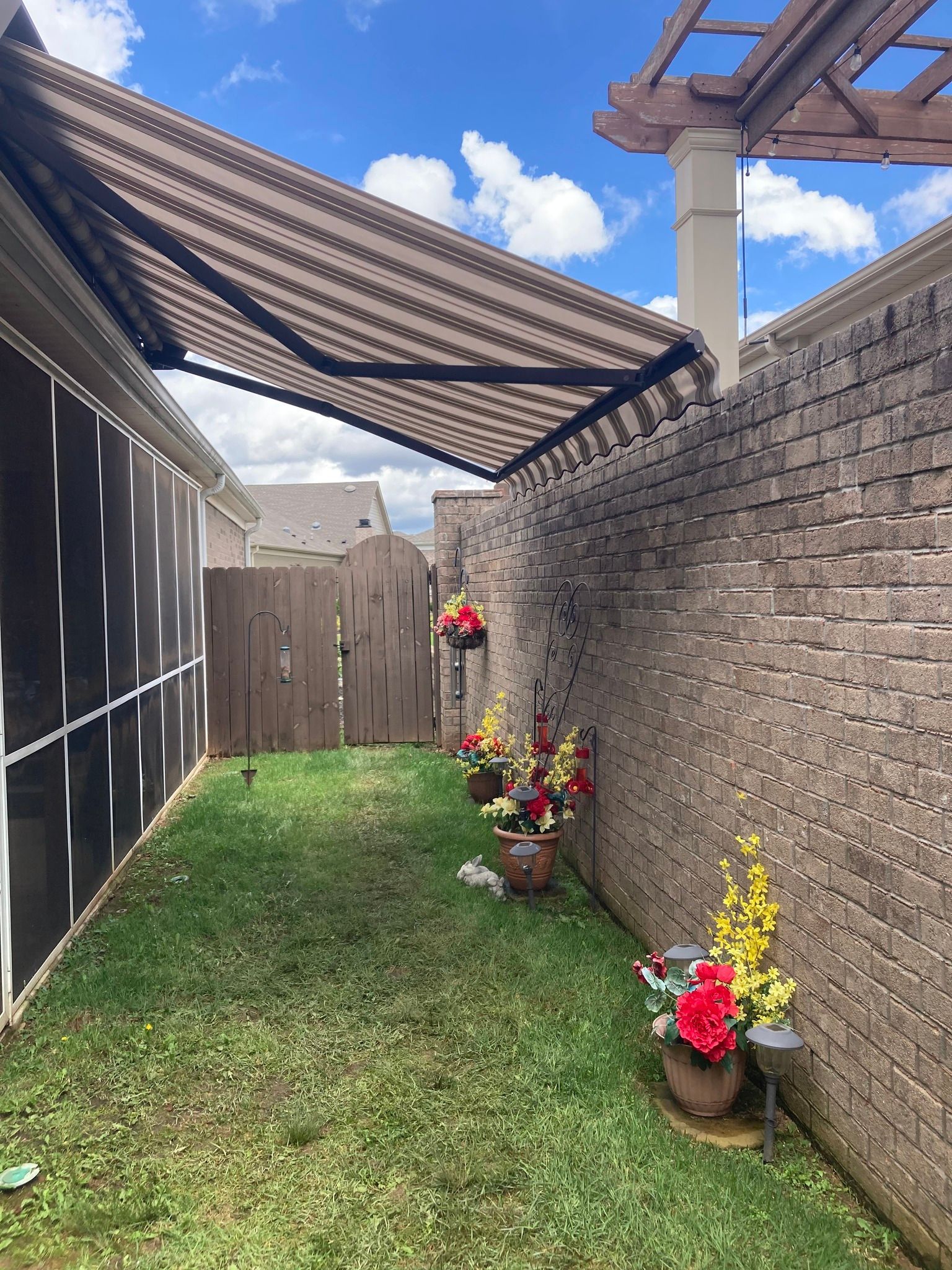 Small backyard with awning, brick wall, fence, and flower pots; sunny day.