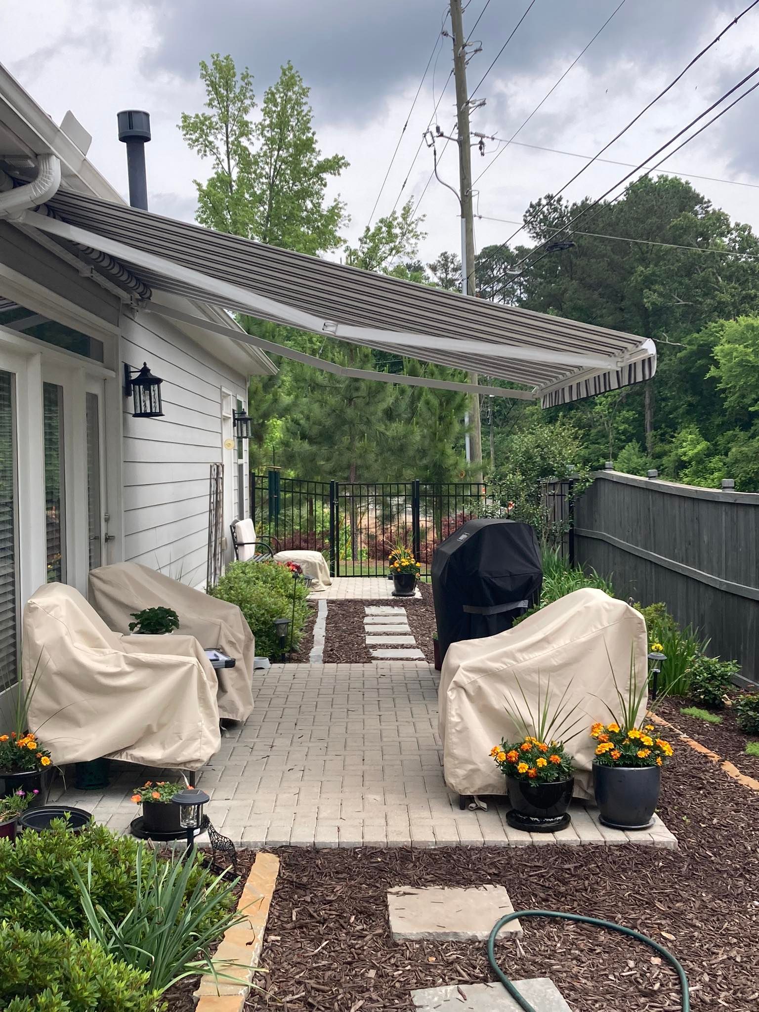 Patio with awning, covered furniture, brick path, landscaping. Cloudy sky.