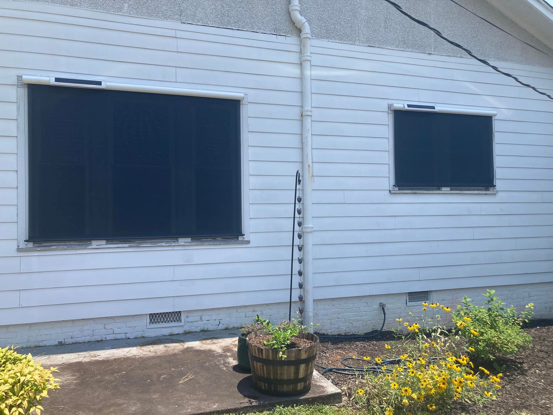 White house with two dark window shades, a plant in a wooden pot, and a drainpipe.