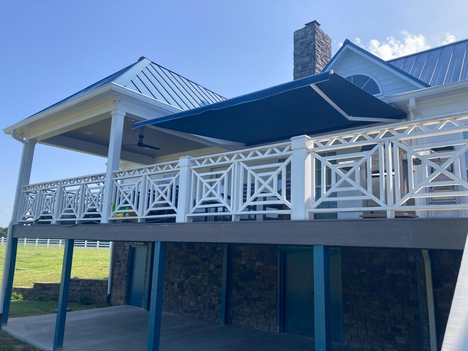 Blue awning over a white-railed deck attached to a light yellow house with a blue roof, under a blue sky.