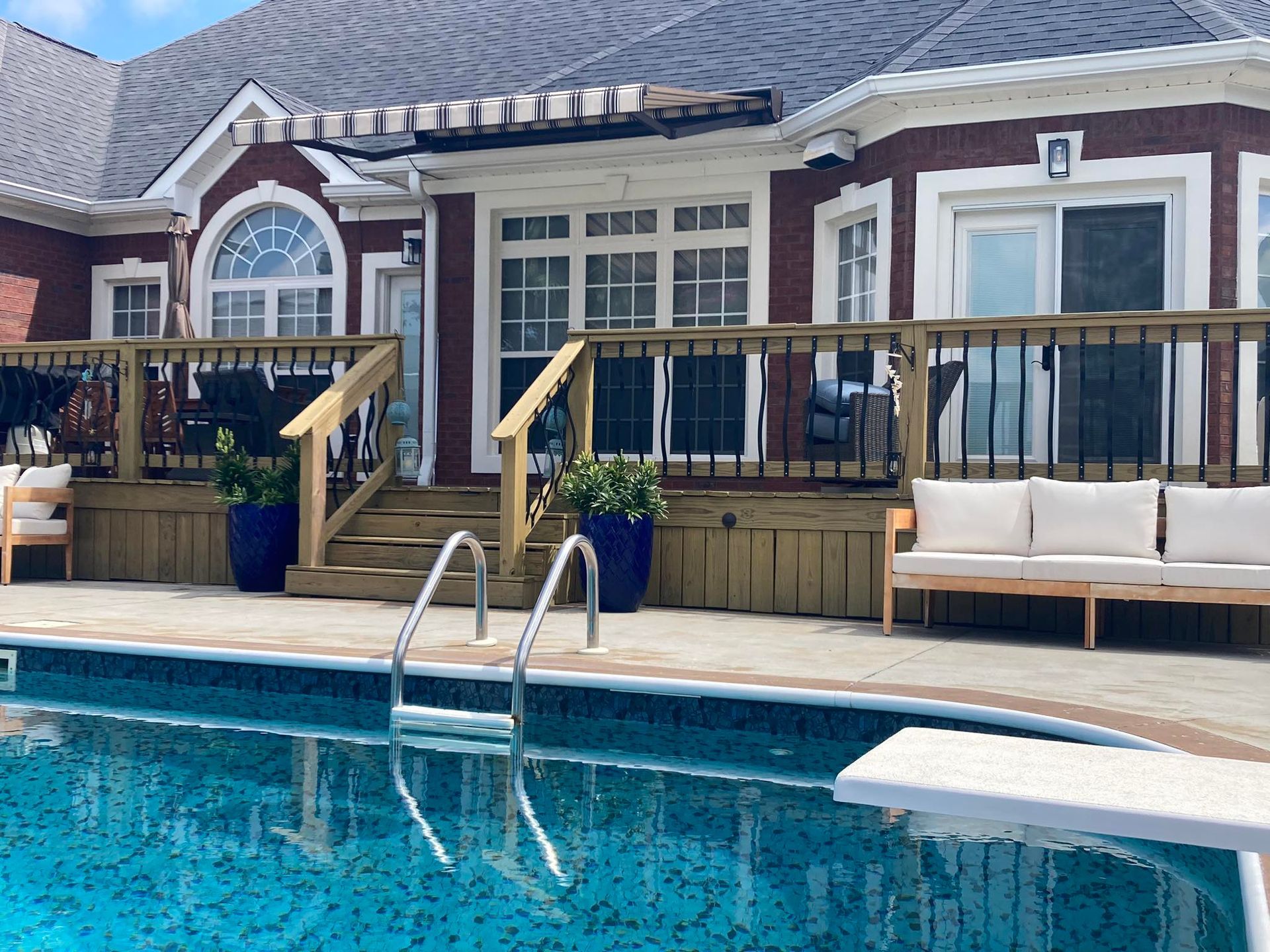 Backyard pool with a wooden deck, steps, and house with brick exterior and white windows.