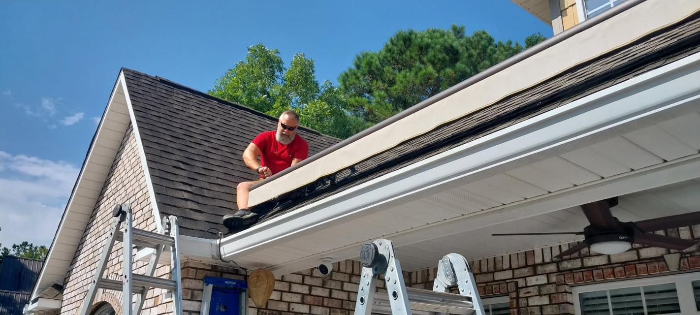 A person on a roof working near a gutter, two ladders are in view, blue sky, brick wall.