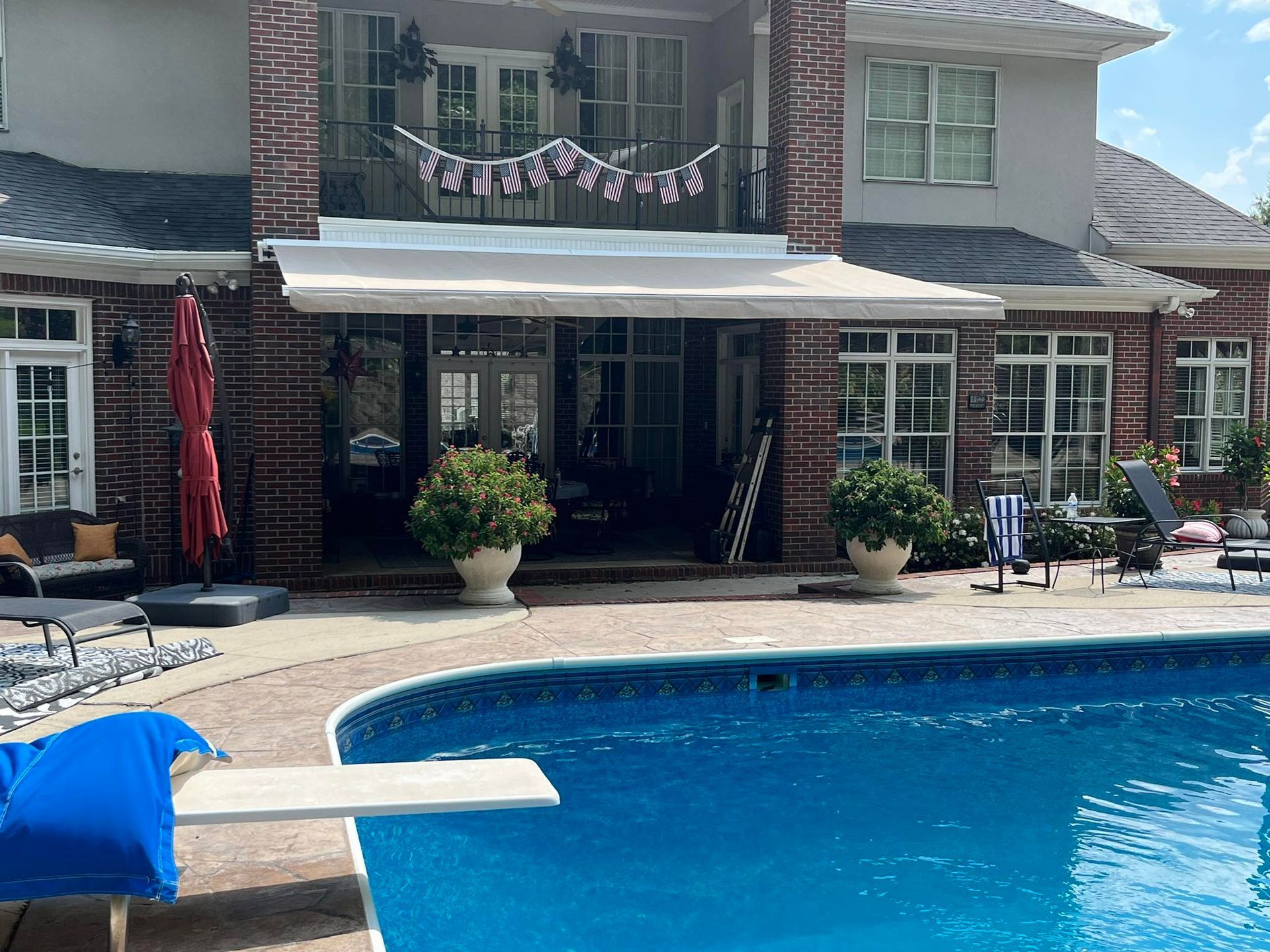 Backyard with a pool; house has a patio awning. Poolside furniture and potted plants are visible. Sunny day.