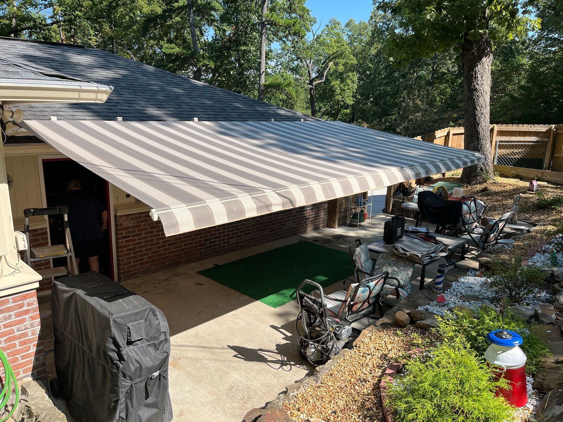 Striped awning extending over a patio. The patio has various items, including a grill and pressure washer.