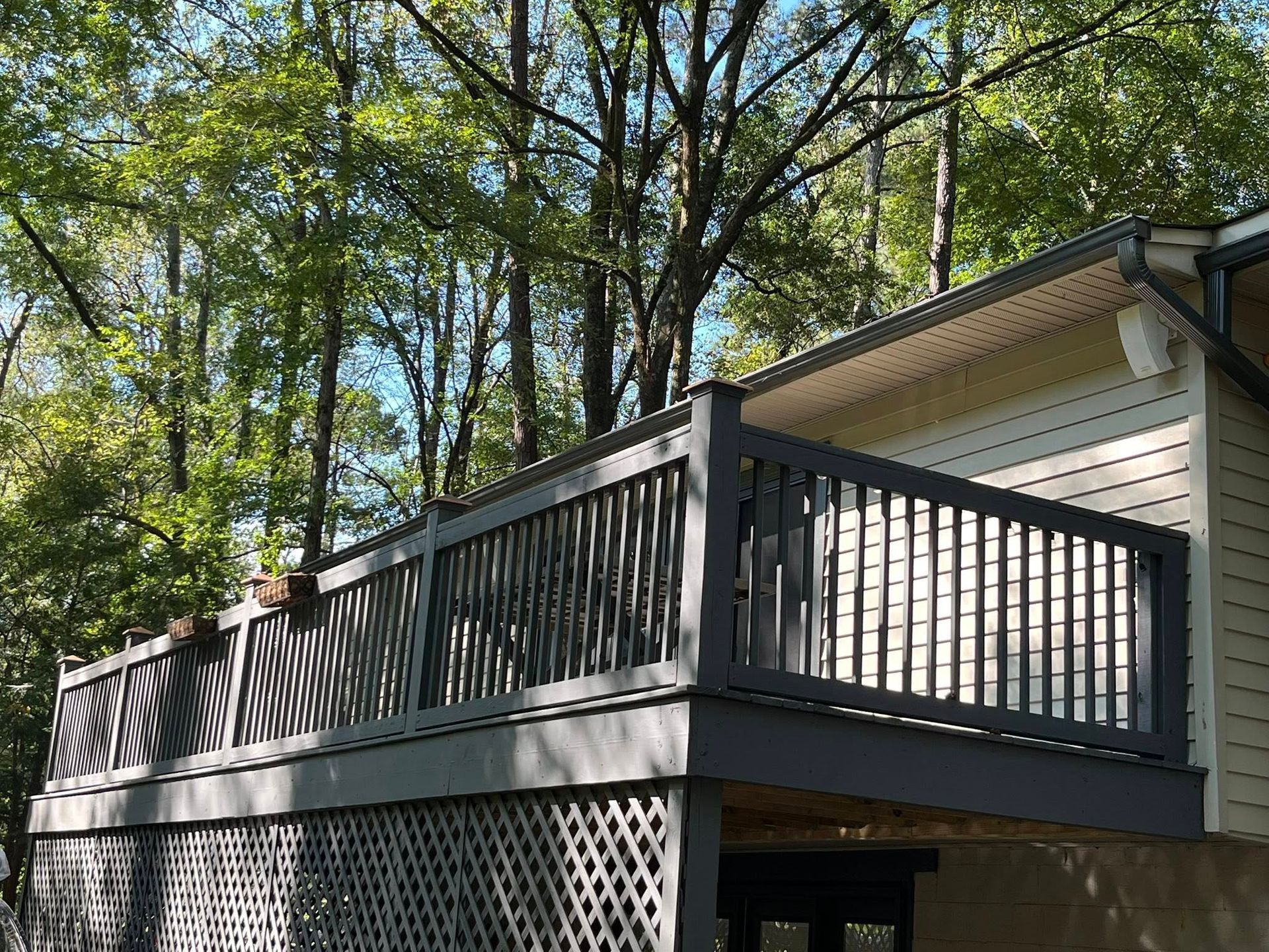 A gray wooden deck with lattice railing, attached to a house with trees in the background.