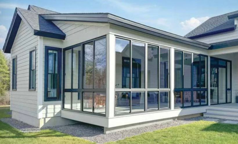 Sunroom addition with glass walls, attached to a light-colored house. Black window frames, green grass, blue sky.