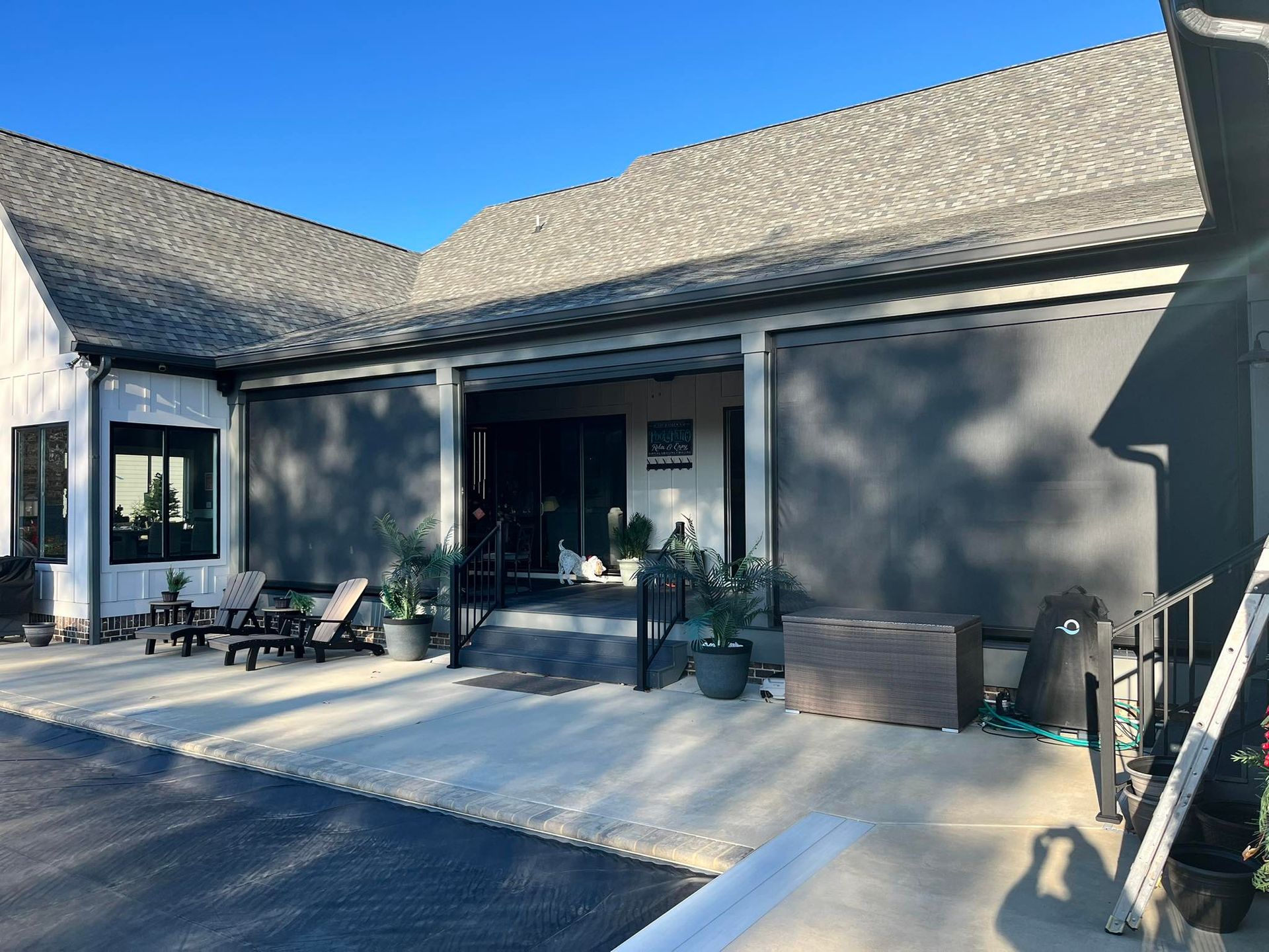 Exterior view of a house with retractable grey sun shades. Patio furniture and plants are visible on the patio.