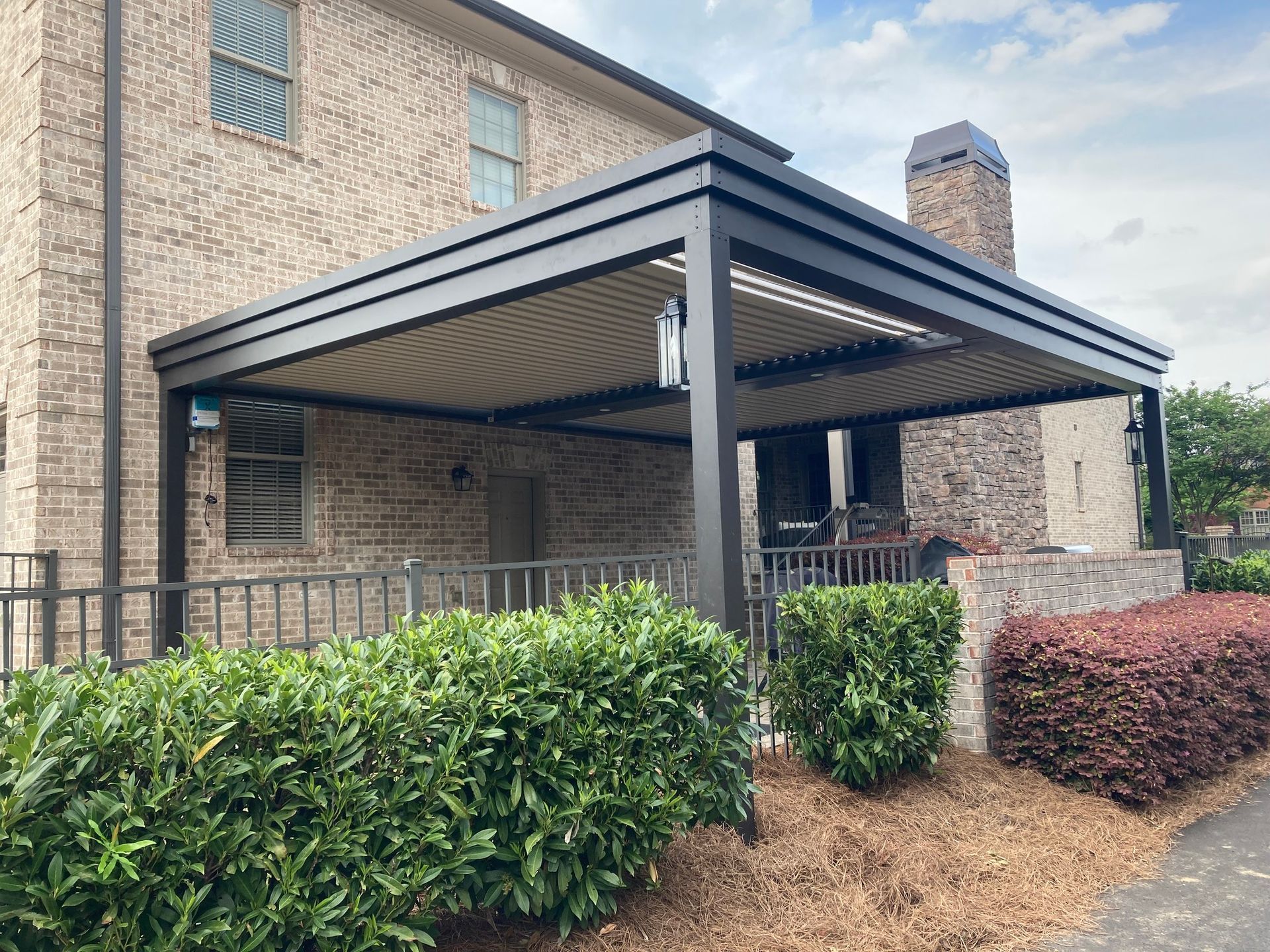 Black pergola attached to a brick house, over a patio with plants and a fence.