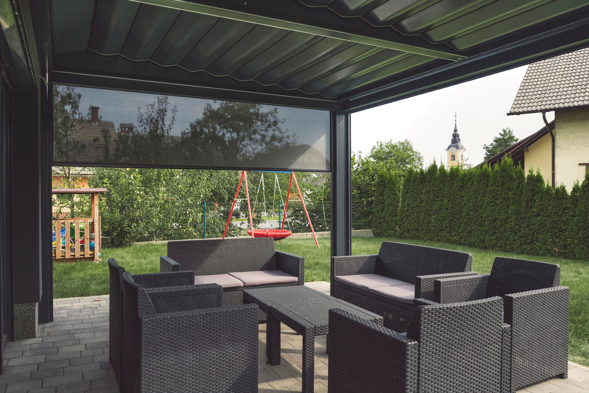 Patio with black wicker furniture under a pergola with a sunshade, overlooking a yard with a swing set.