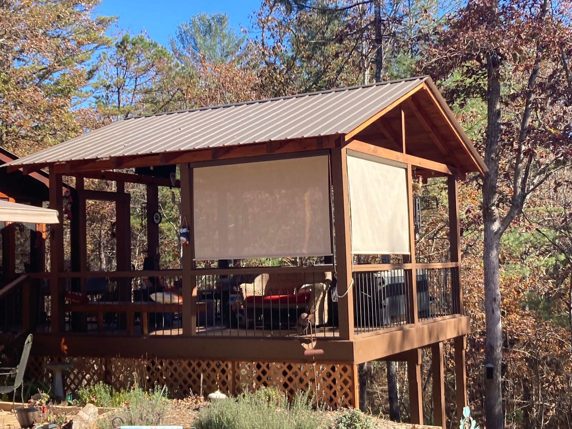 Wooden gazebo on a raised deck with shades, overlooking a wooded area. Brown, beige, and green colors.
