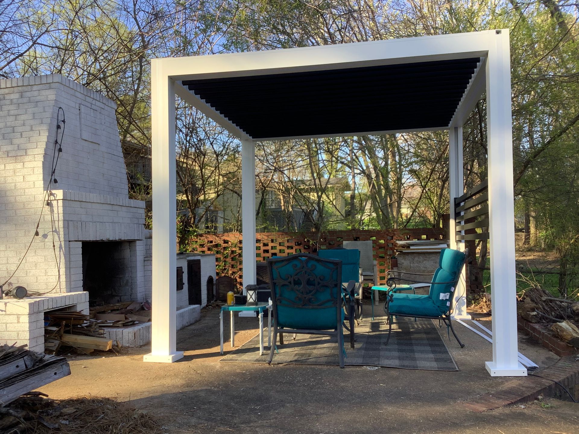 White pergola with teal patio furniture on a concrete patio, with a brick fireplace in the background.