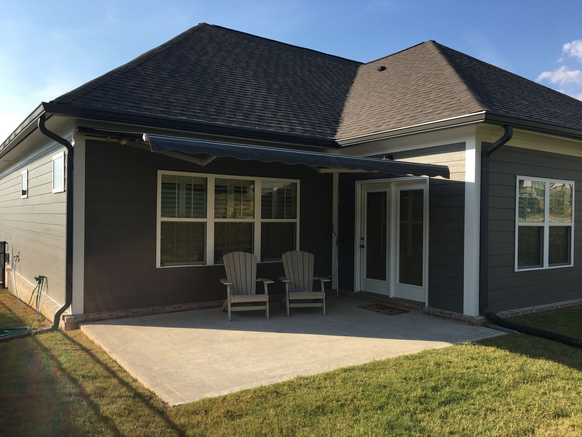 Back patio of a house with two chairs, concrete slab, dark gray siding, and a retractable awning.
