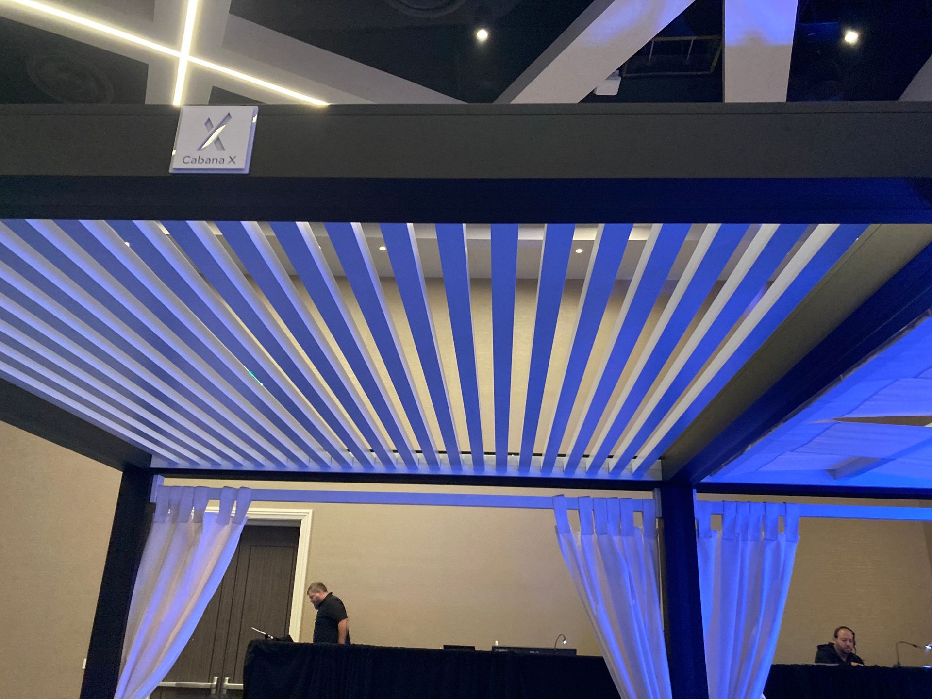 A black pergola with white horizontal slats, white curtains, and people at a table.