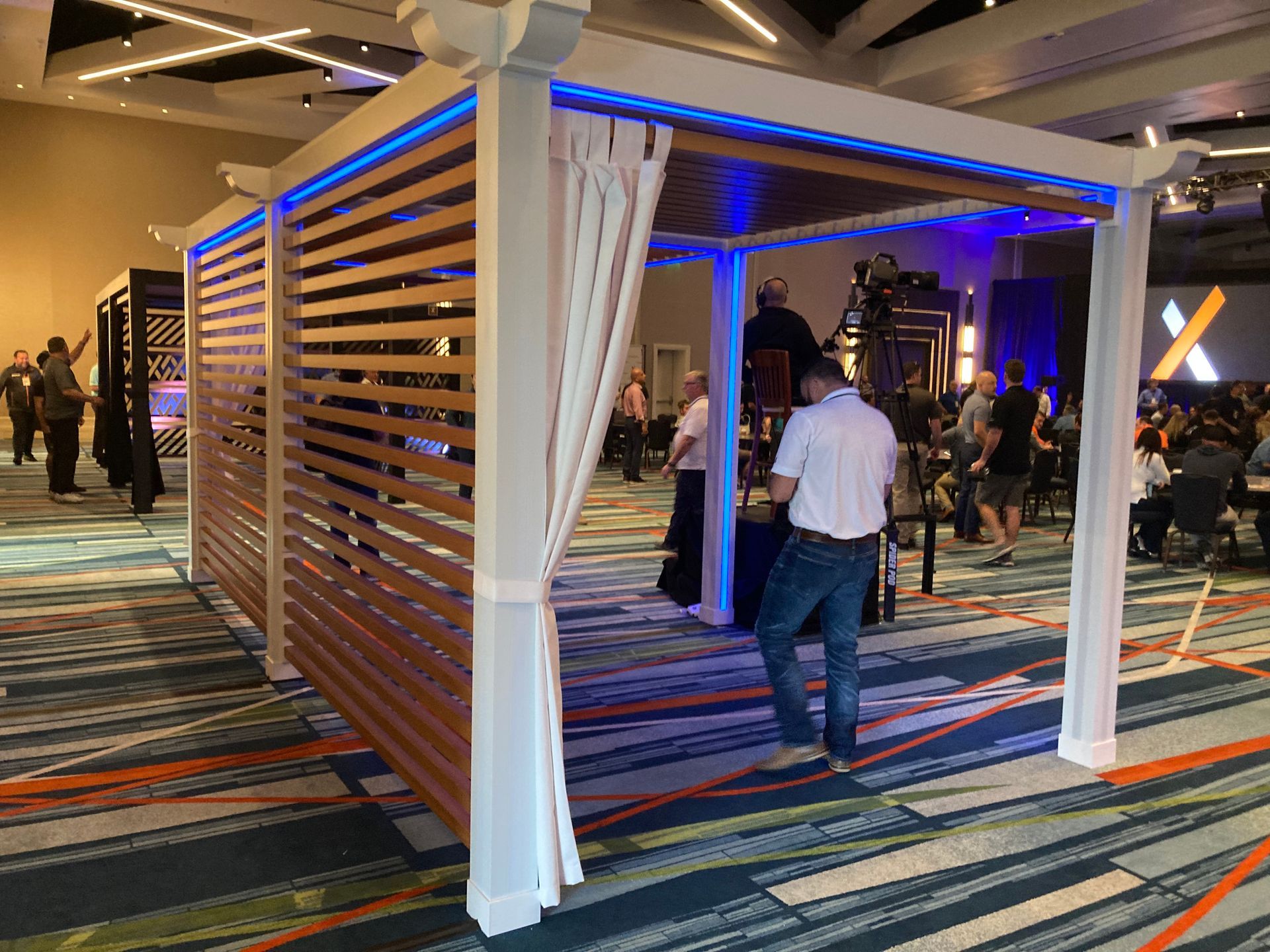 White pergola structure with blue lighting and wooden slats, inside a large event space with people.