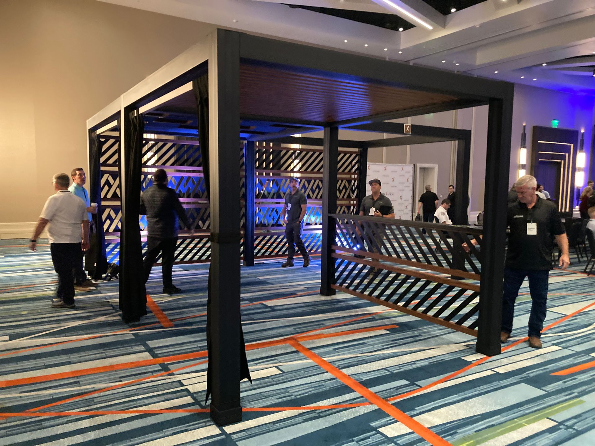 A black metal pergola with decorative wooden panels, in a convention hall, people milling around.