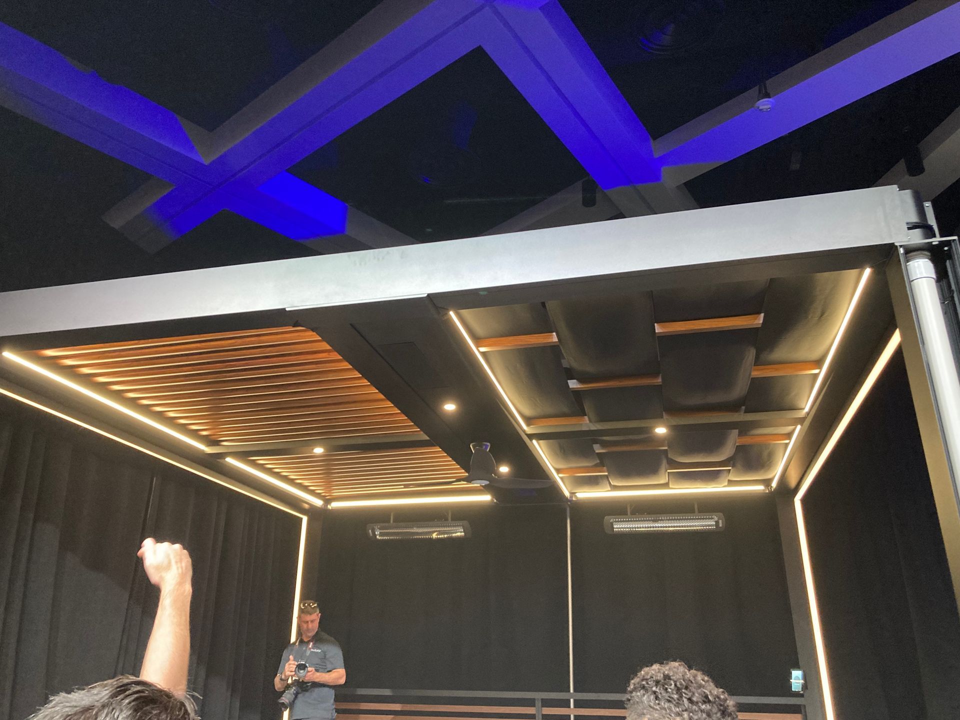 A speaker on a stage in a dark room. The stage has a wooden ceiling with lighting. Blue lights are on the ceiling above.