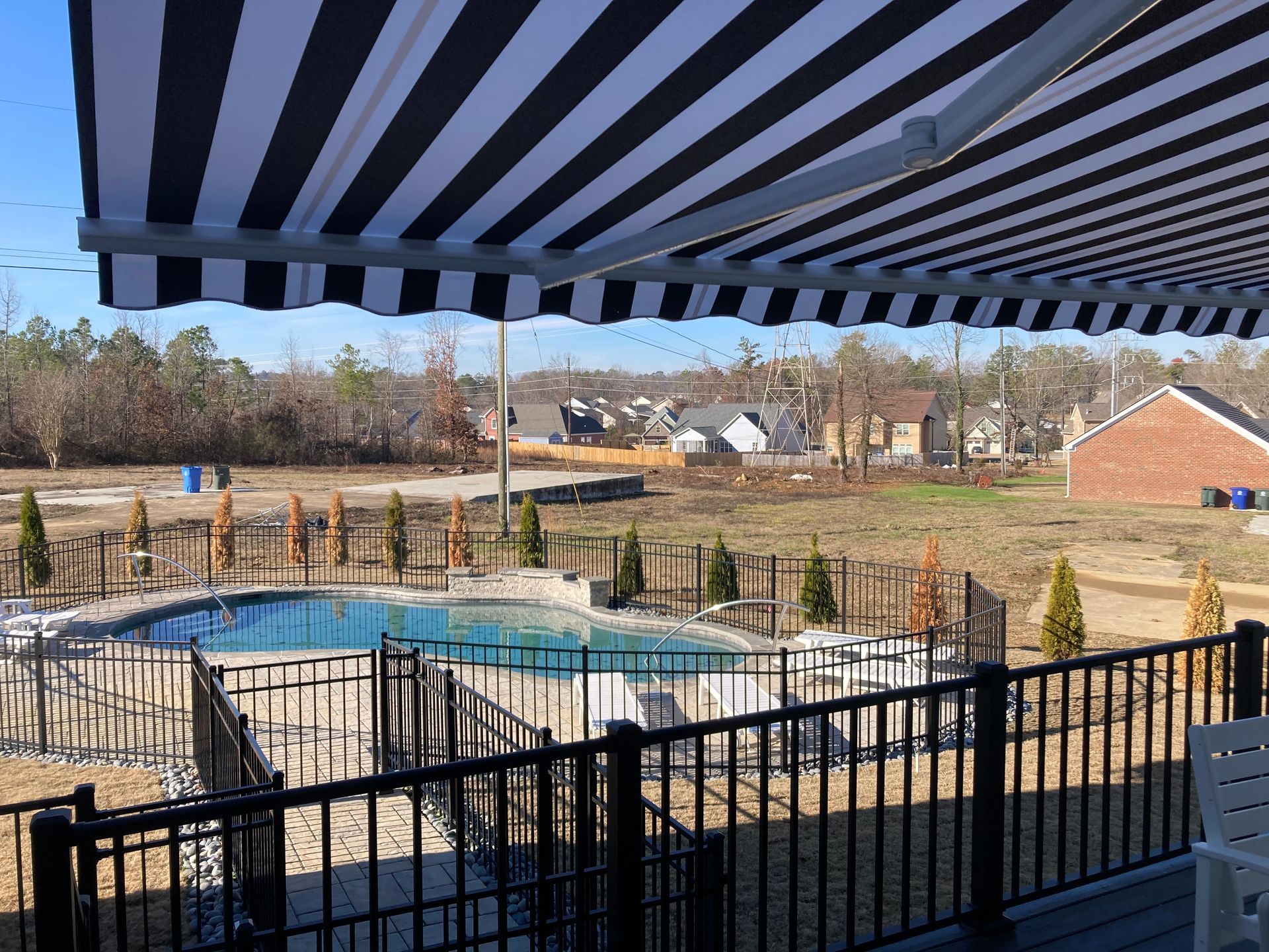 Pool area with black and white striped awning and black fence; houses in background.