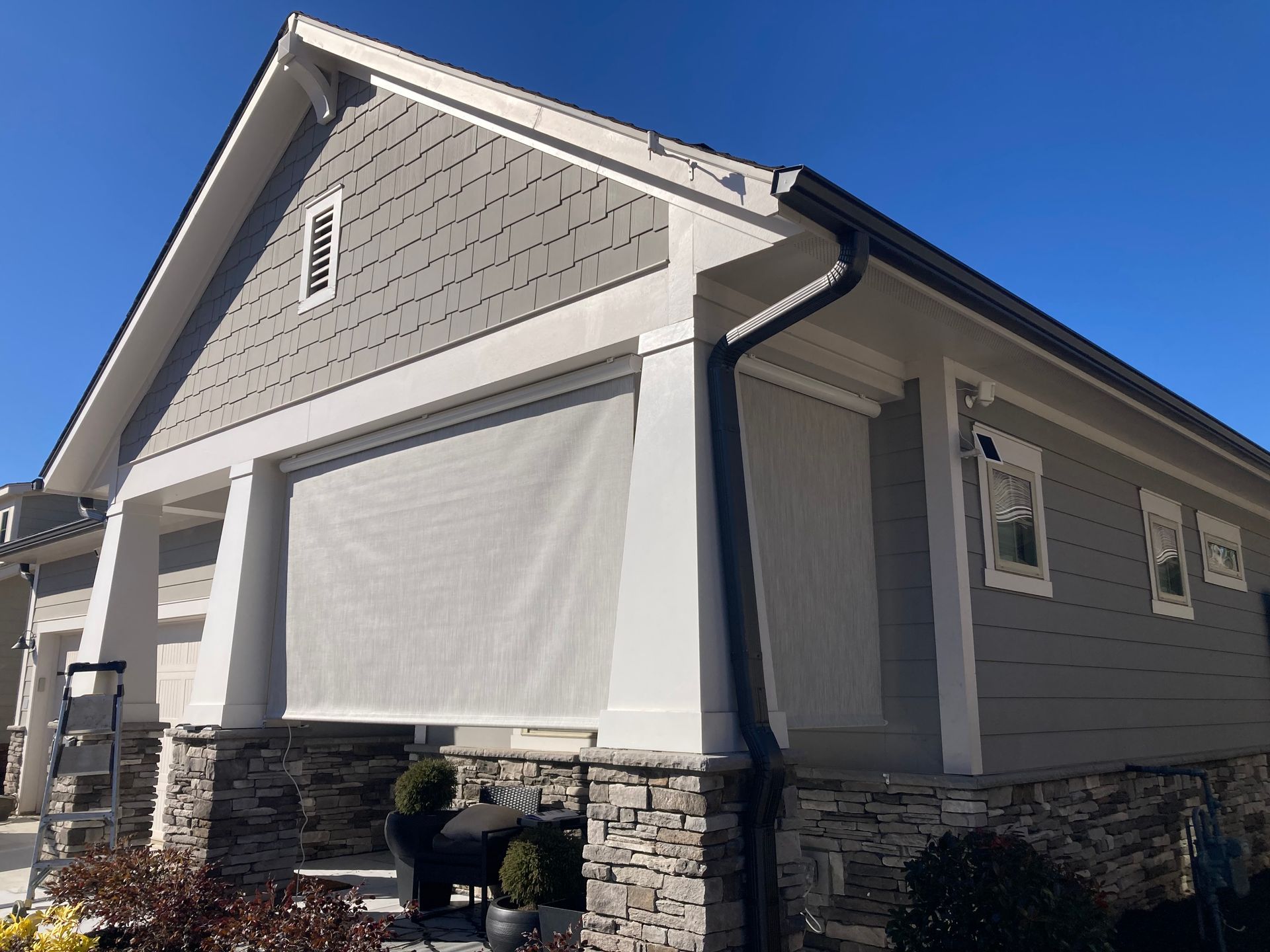A house exterior with retractable screen shade. Gray siding, white trim, stone base, and blue sky.