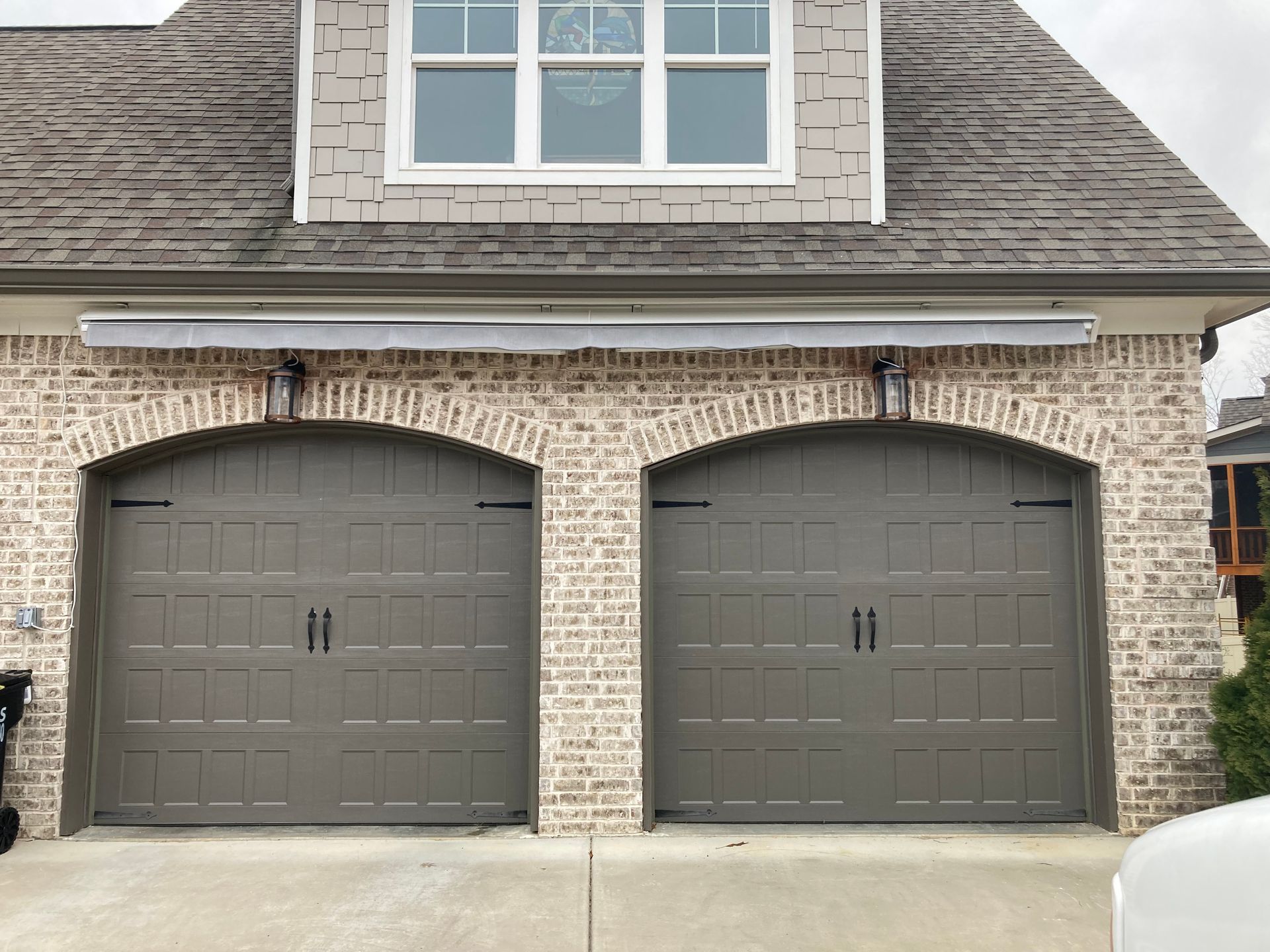 Two gray garage doors under a brick facade with an awning, topped by a window and a gray roof.