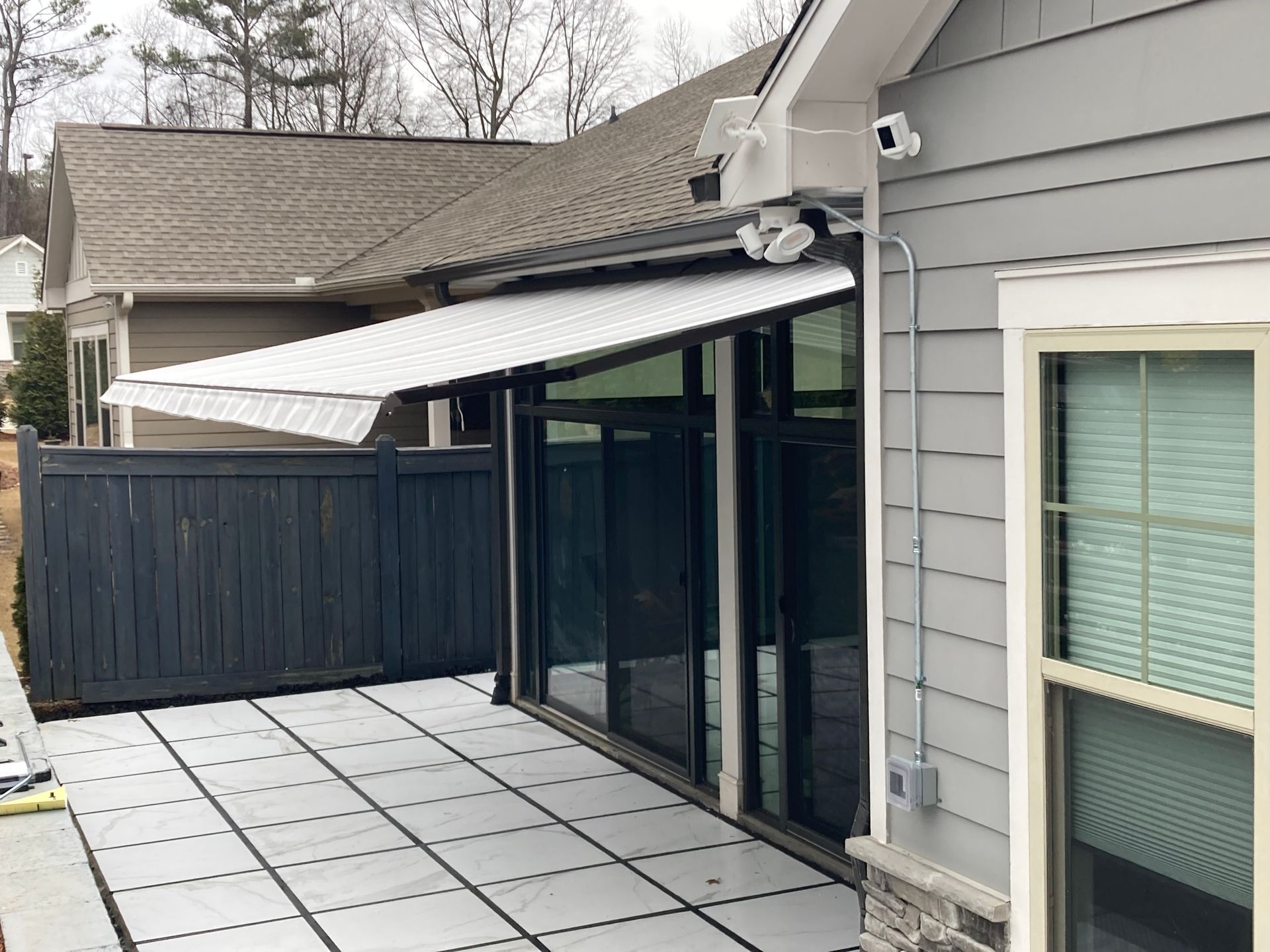 A white retractable awning extends over a patio.  The patio has gray tiles,  glass doors, and a dark wooden fence.