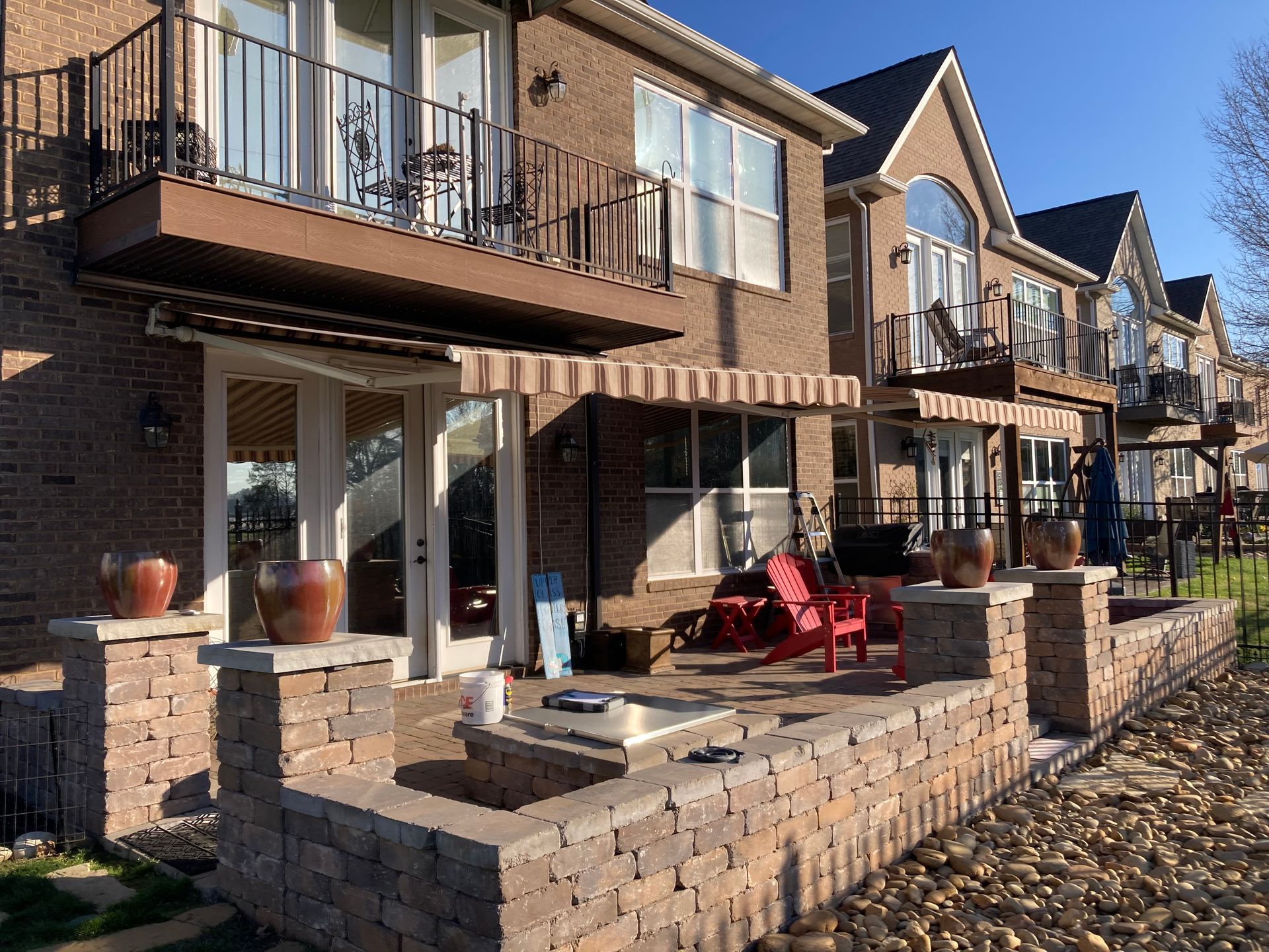 Brick townhouses with balconies and awnings. Patio area with a low wall, red chairs, and potted plants.