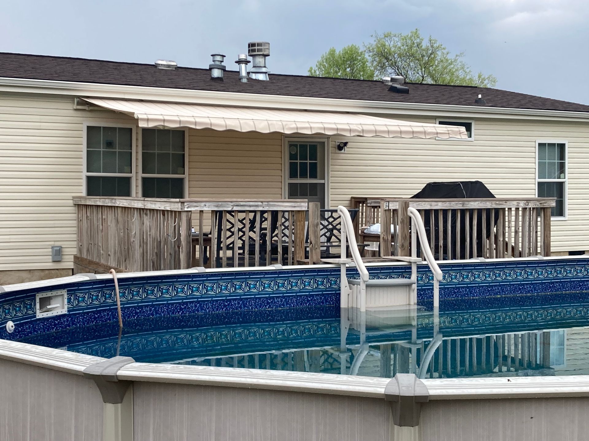 Backyard pool with a wooden deck, retractable awning, and a house in the background.
