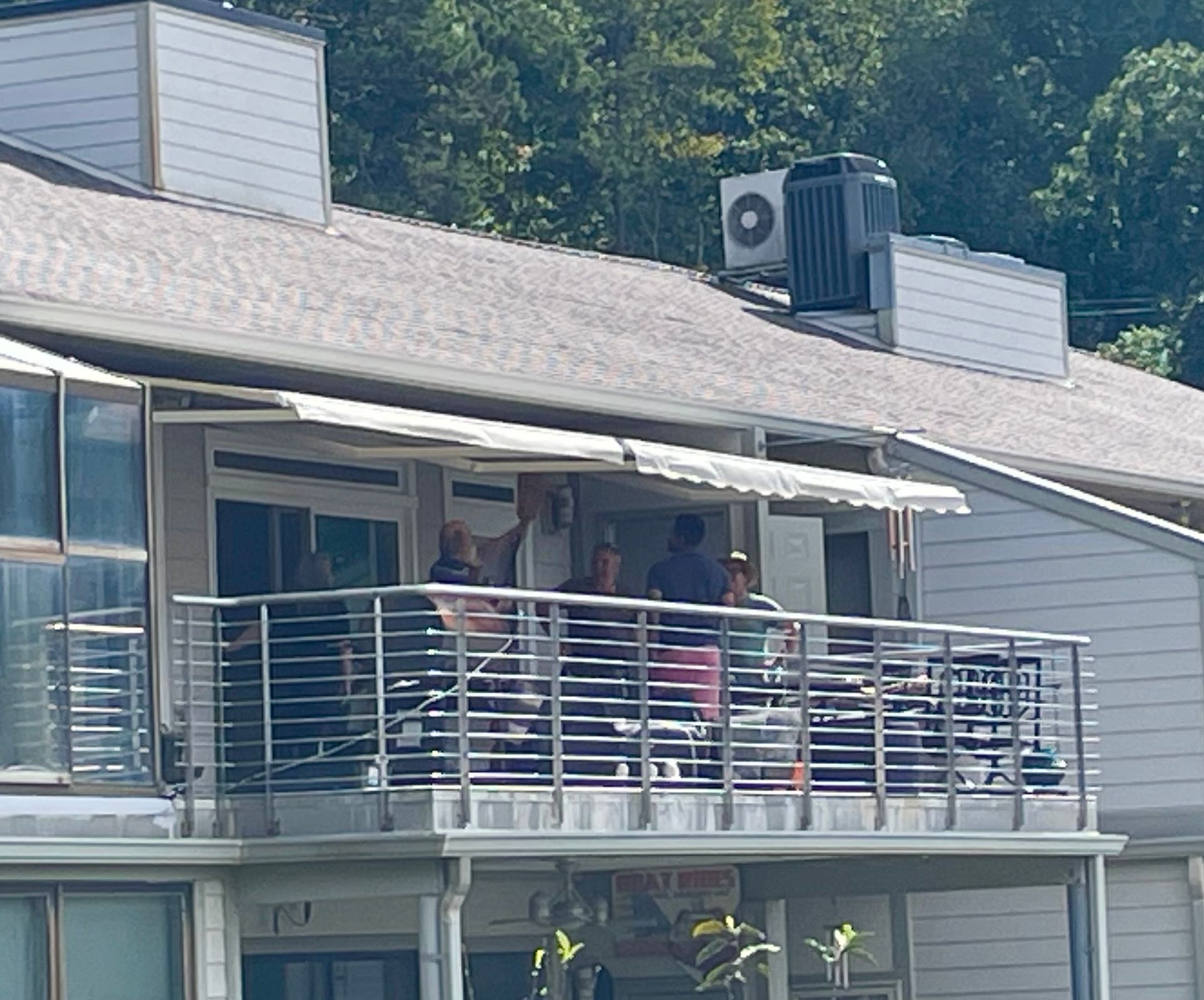People on a balcony, under an awning. Buildings are light-colored with trees in the background.