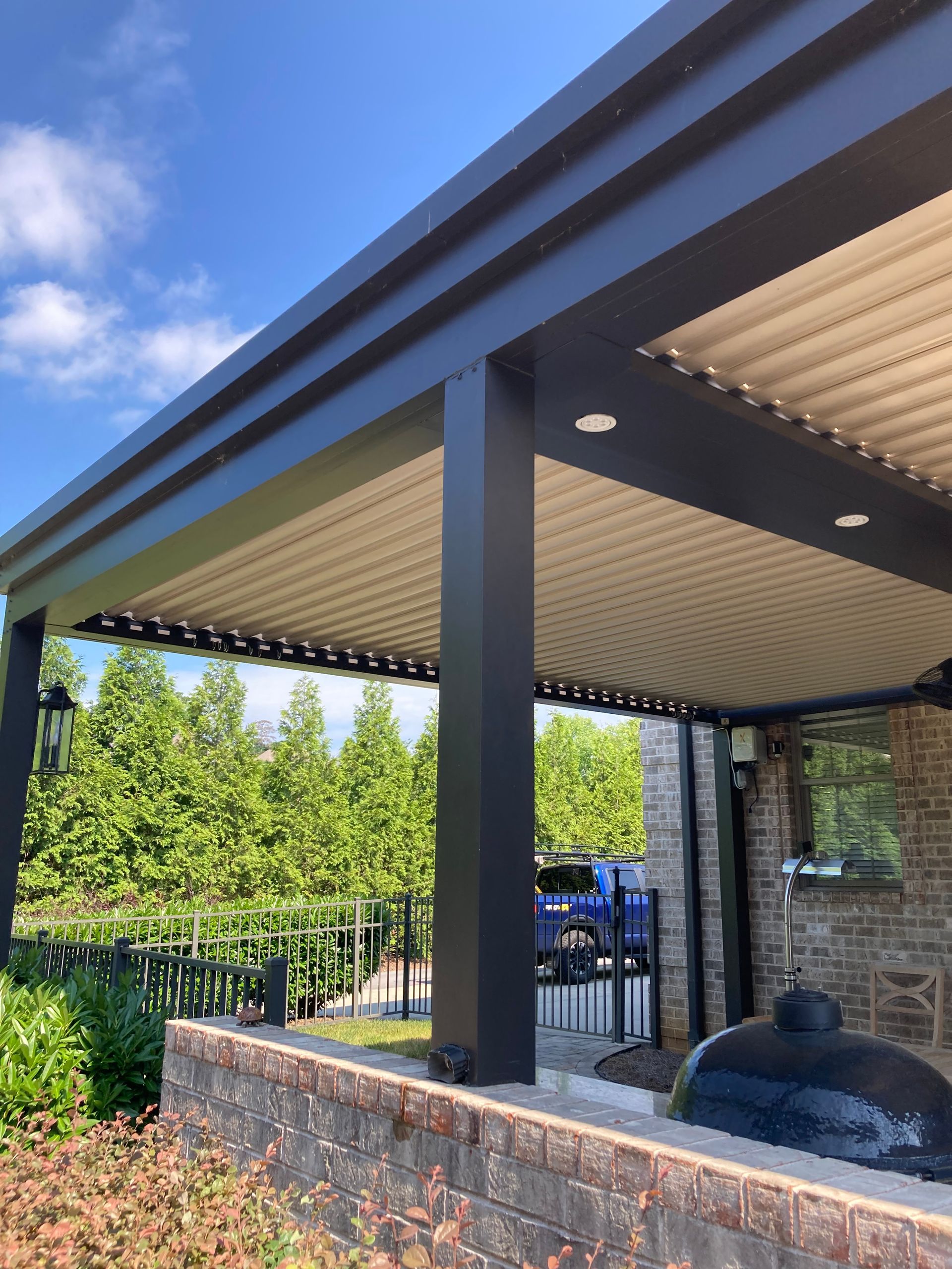 Outdoor patio with a black pergola, brick wall, and green foliage under a blue sky.
