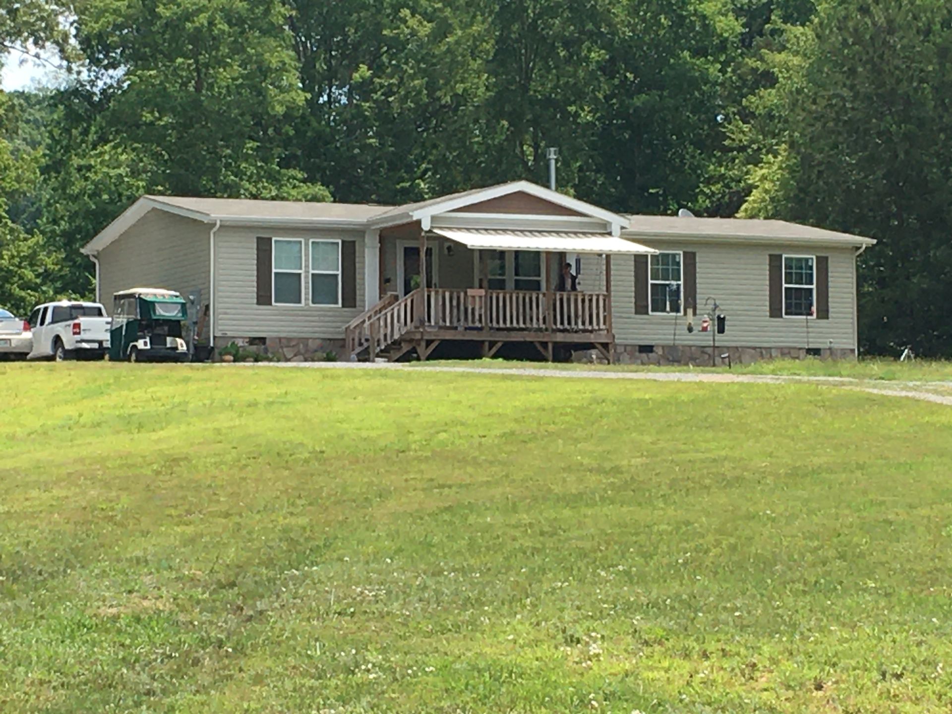 Ranch-style house with tan siding, brown shutters, and small porch. Cars are parked to the left, green lawn in front.