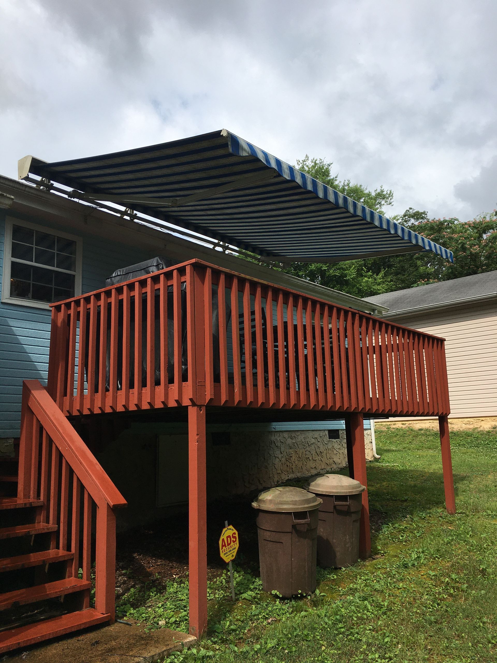 Wooden deck with red railing and blue/white striped awning attached to a blue house. Two trash cans.