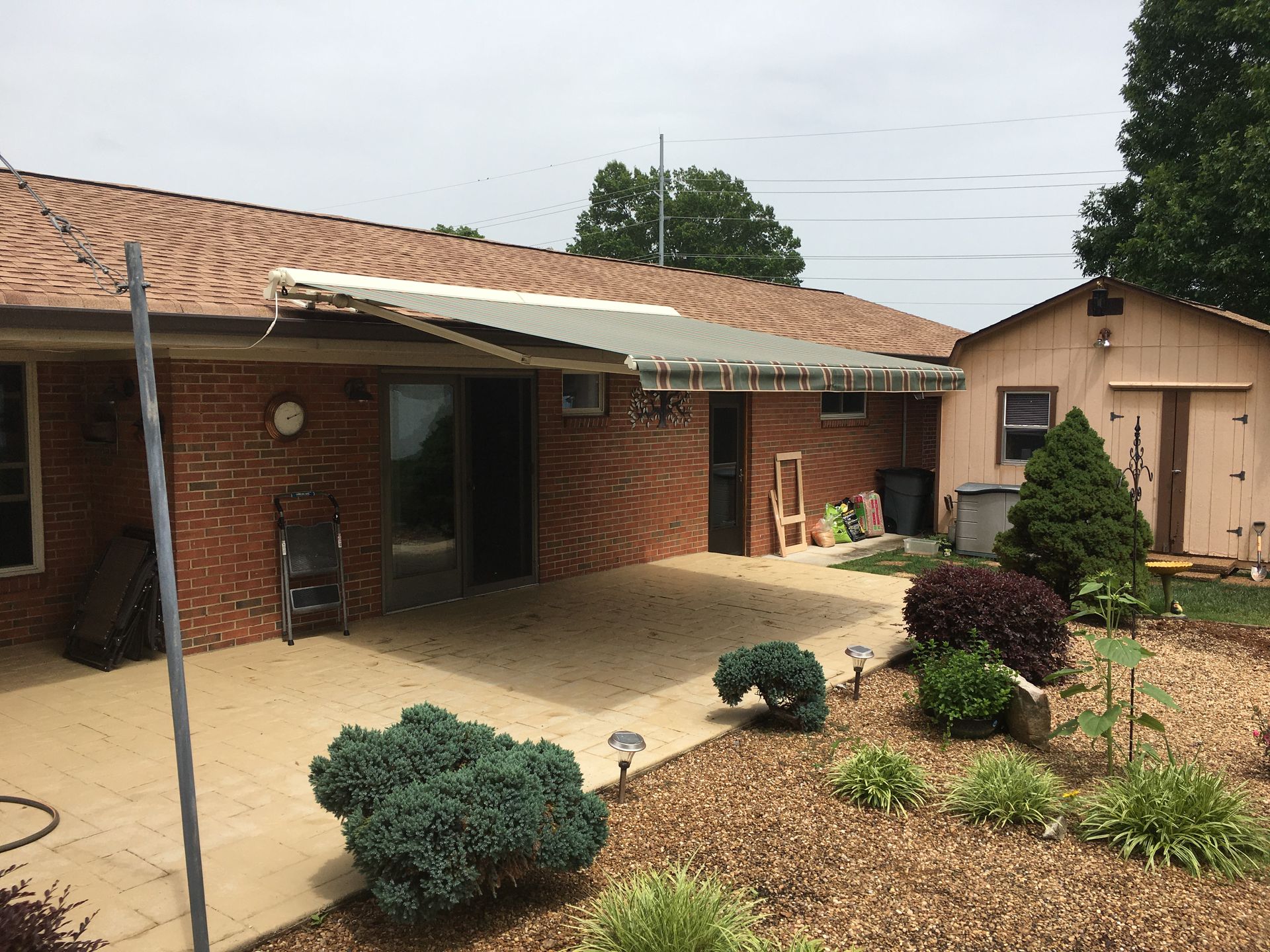 Backyard with brick house, damaged awning, concrete patio, shed, and landscaping.