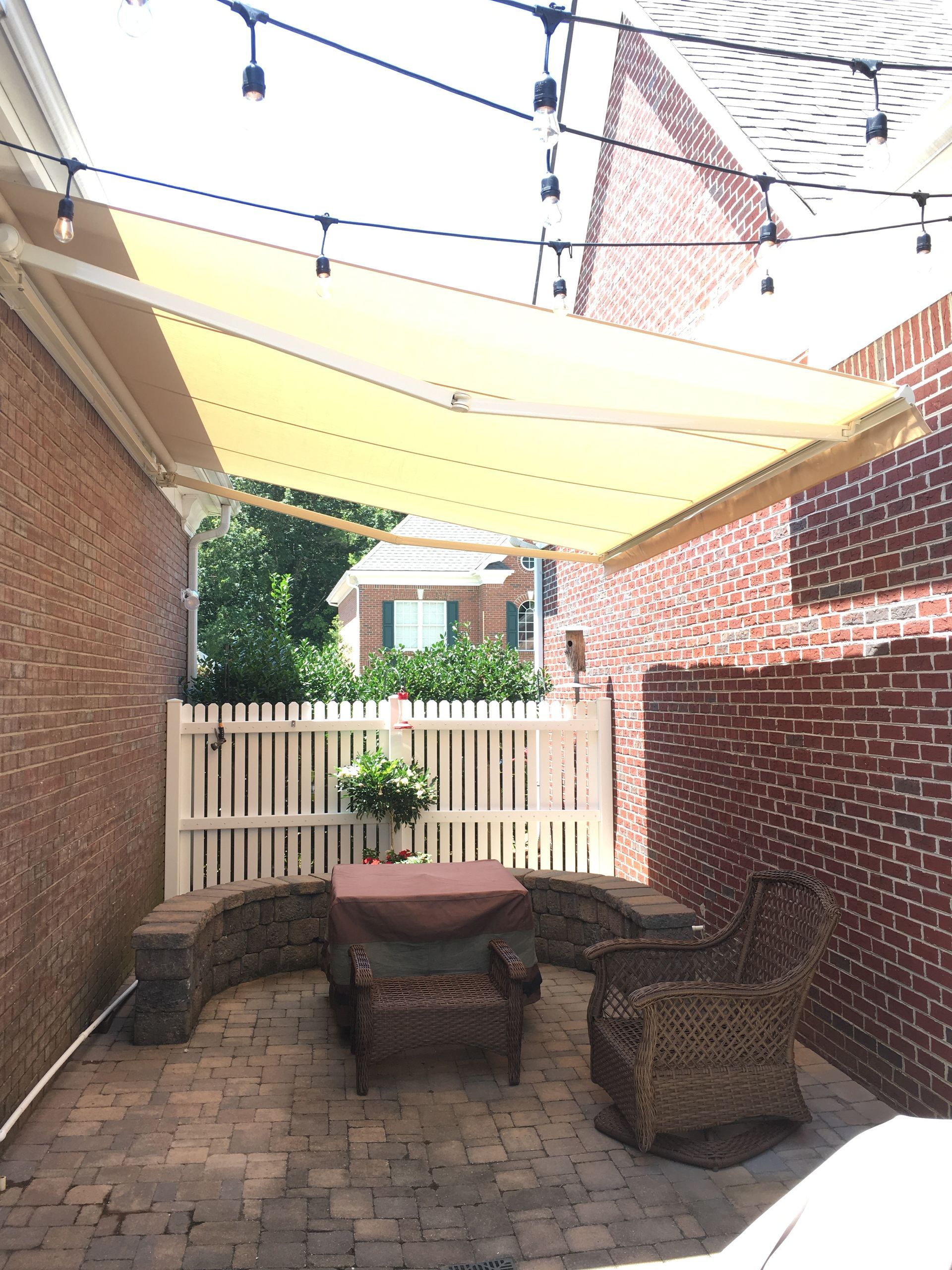 Patio with brick walls, tan awning, string lights, white fence, seating, and plants.
