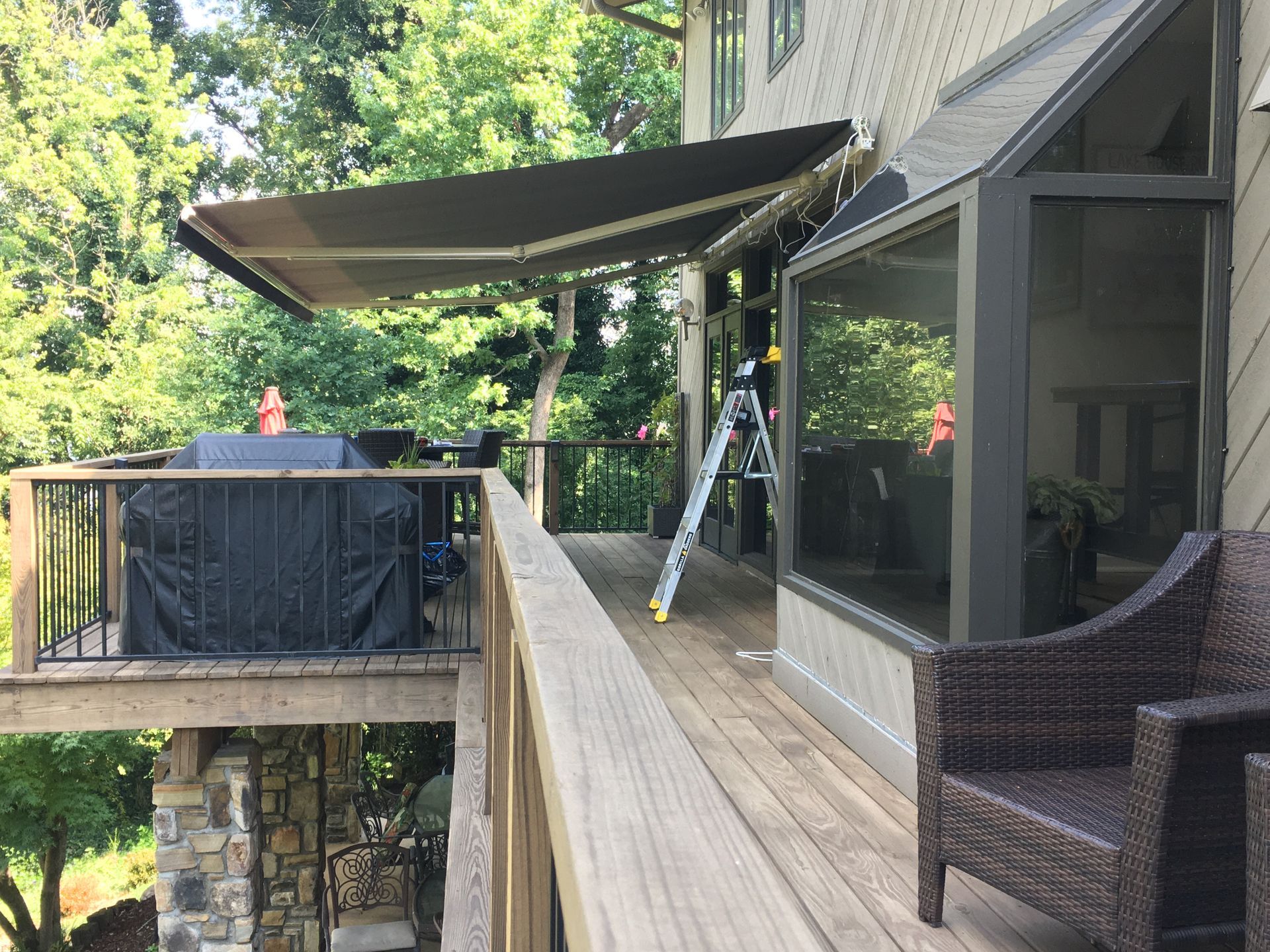 Deck with retractable awning extended; grill covered; two-story house with bay window and ladder; trees in background.