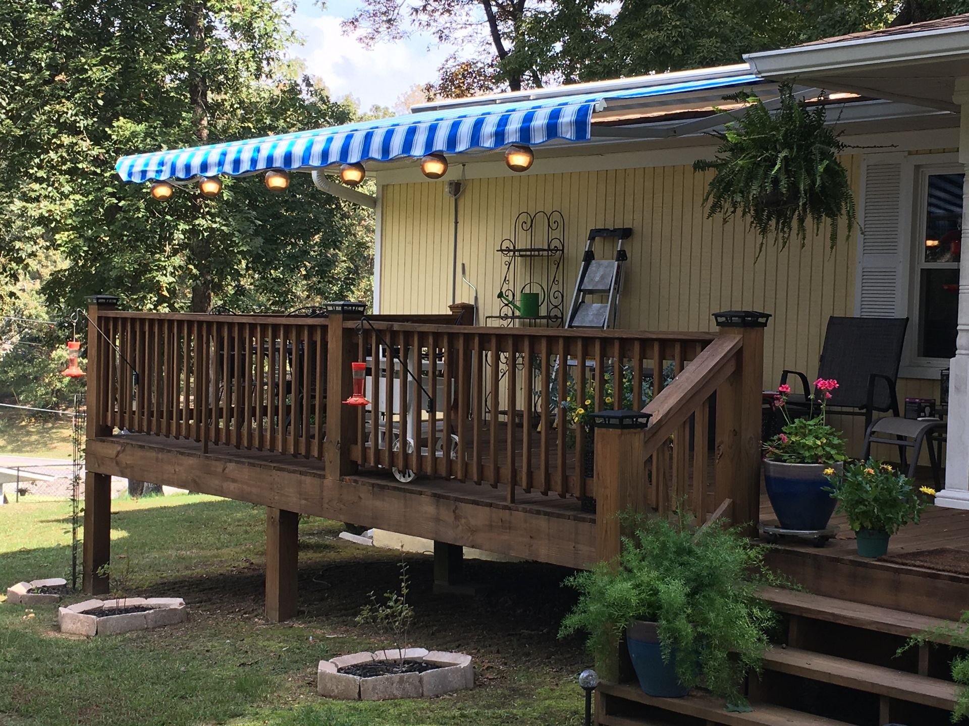 Wooden deck with blue and white striped awning and string lights attached to a yellow house.