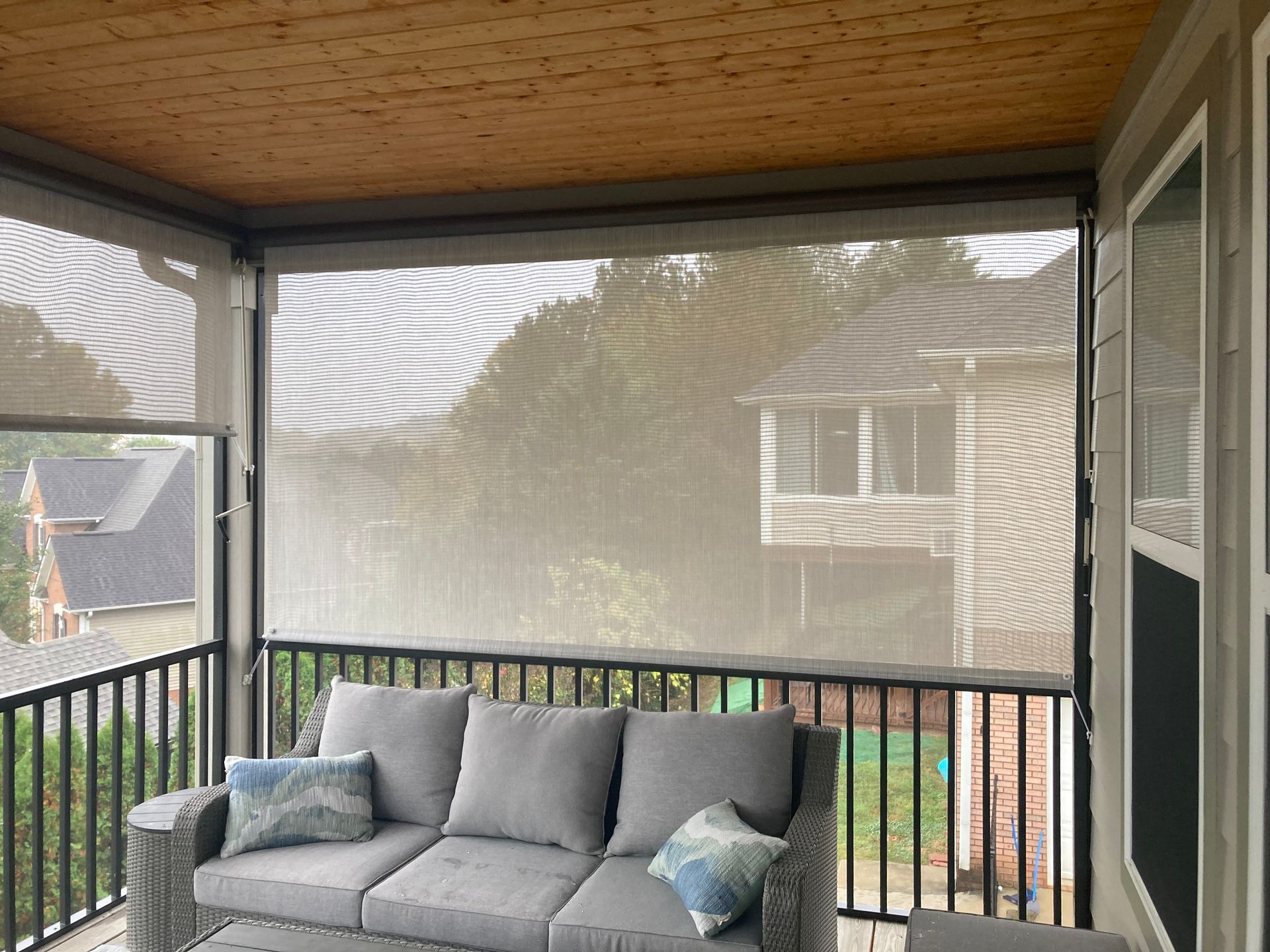 Outdoor patio with beige sun shades, gray couch, and view of trees.
