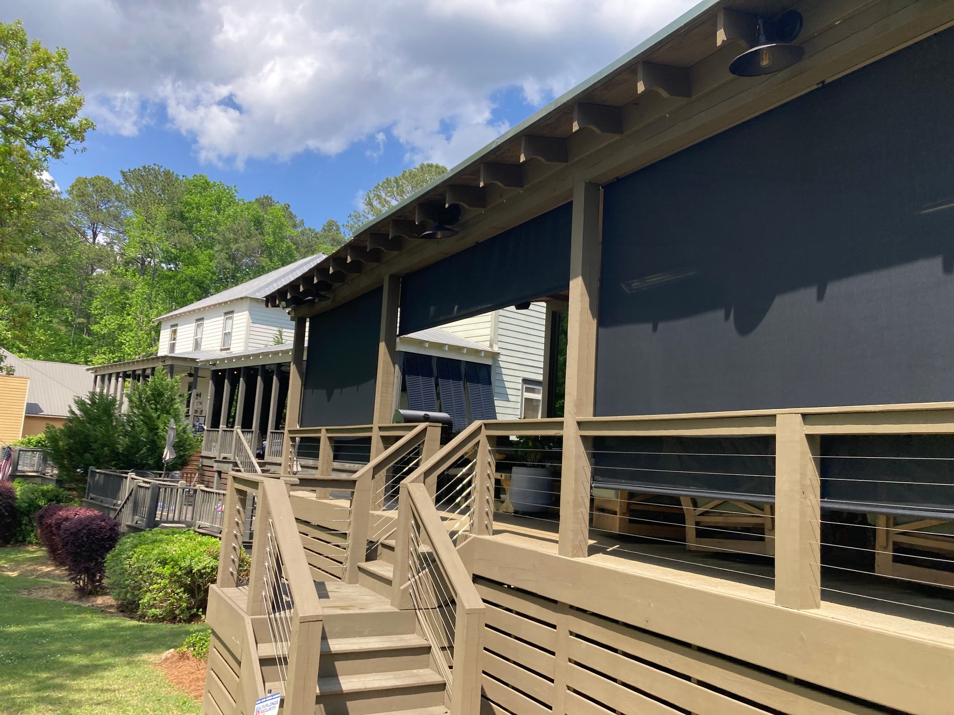 Wooden deck with black shades, leading to a covered outdoor seating area. Green grass and trees in background.