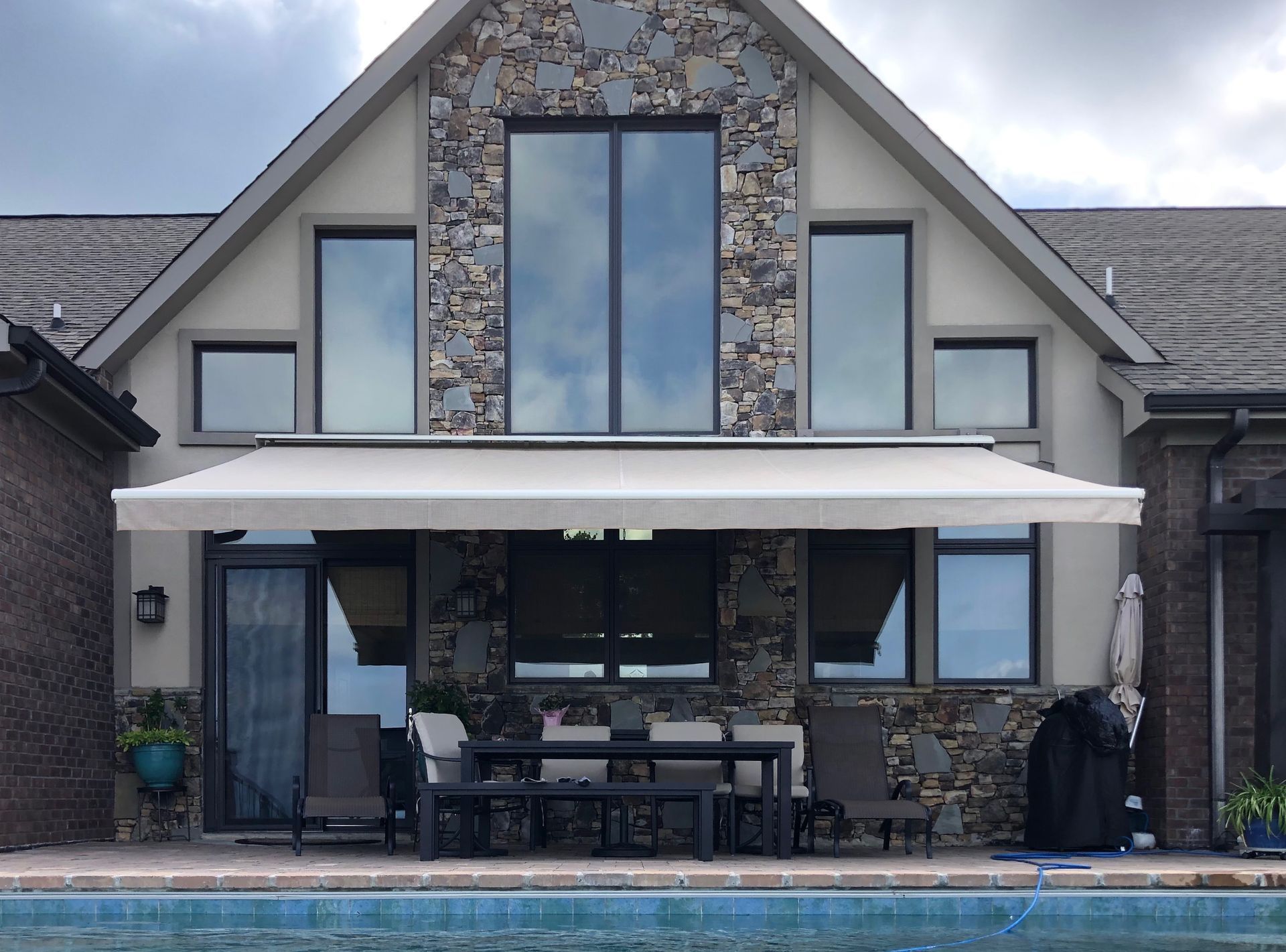 Beige awning over patio with table and chairs, stone facade house, and swimming pool.
