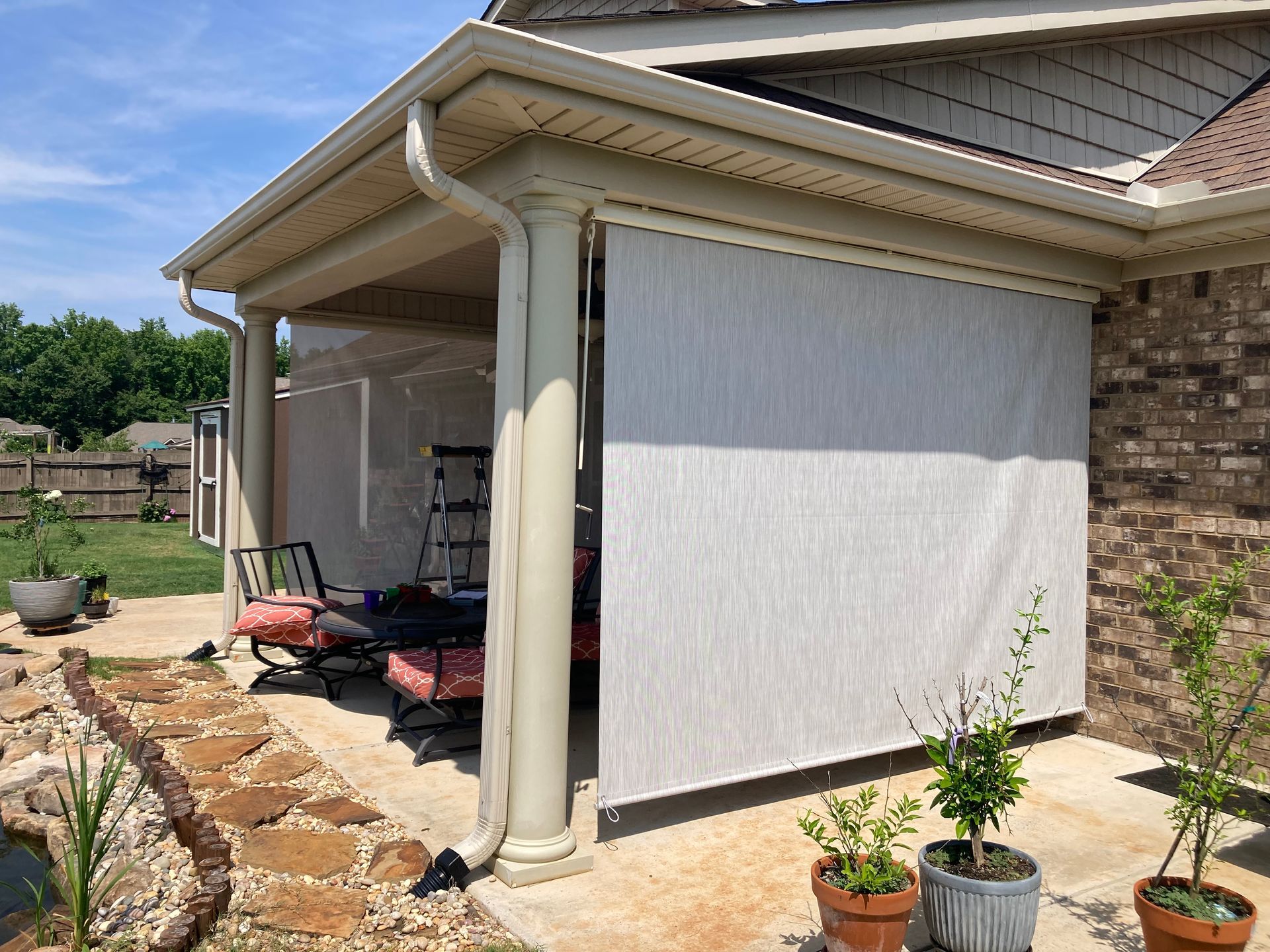 Covered patio with roll-down shade. Beige pillars and roof, light-colored shade, brick wall.