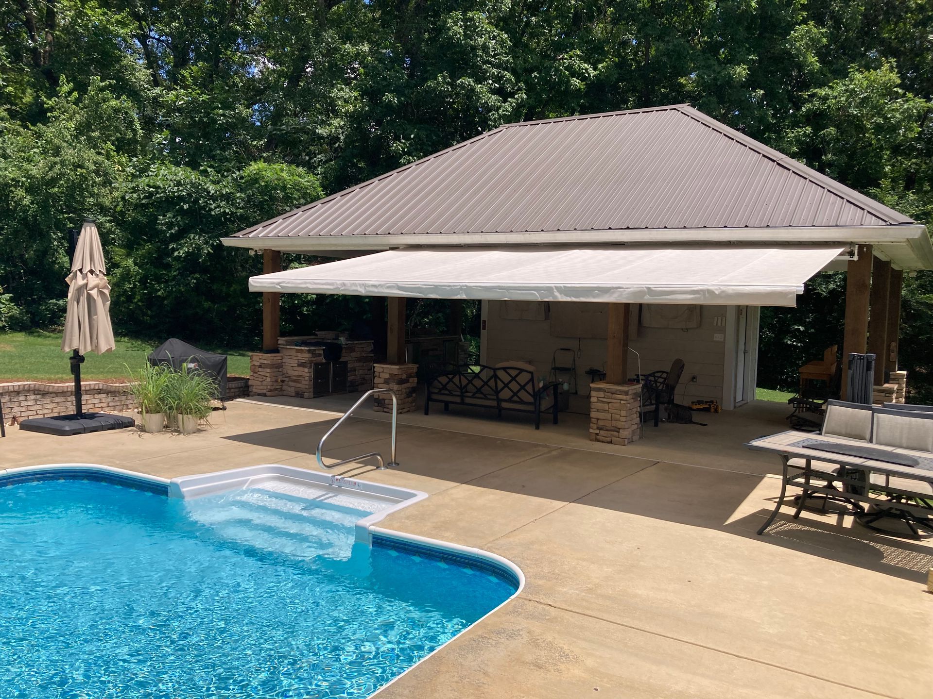 Poolside patio with retractable awning, adjacent to a pool, surrounded by trees.