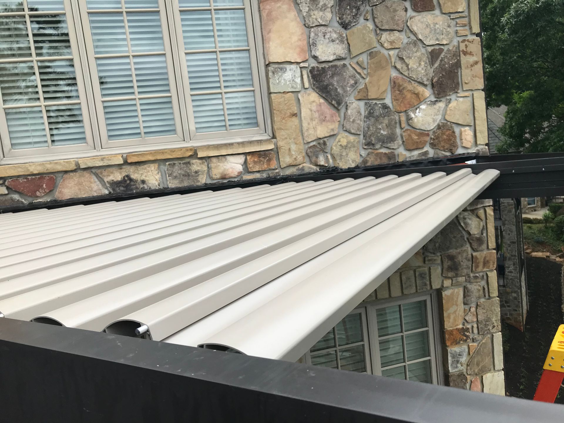 Beige corrugated roof attached to a stone-clad building. Black trim and a window with blinds visible.