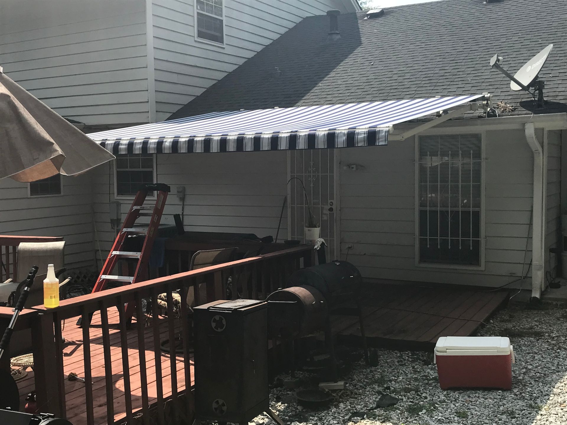 Backyard deck with striped awning, grill, and white house. Red cooler sits on gravel.