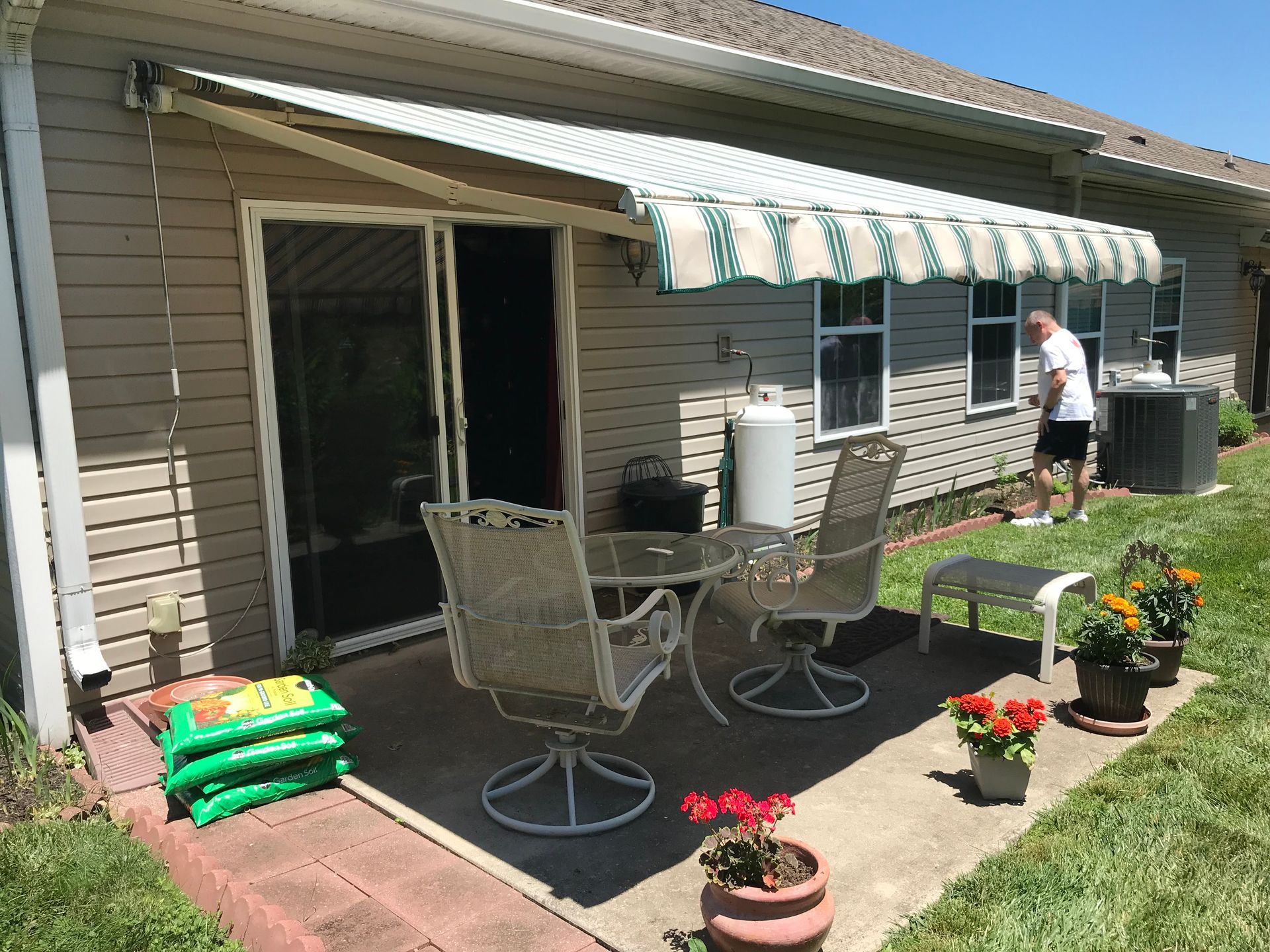 Patio with awning, table, chairs, plants, and a person near a building.