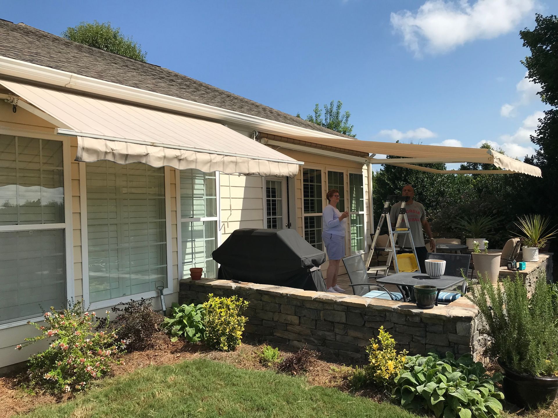 Beige awnings extending from a light-colored house. Two people work on an awning near a stone wall, blue sky.