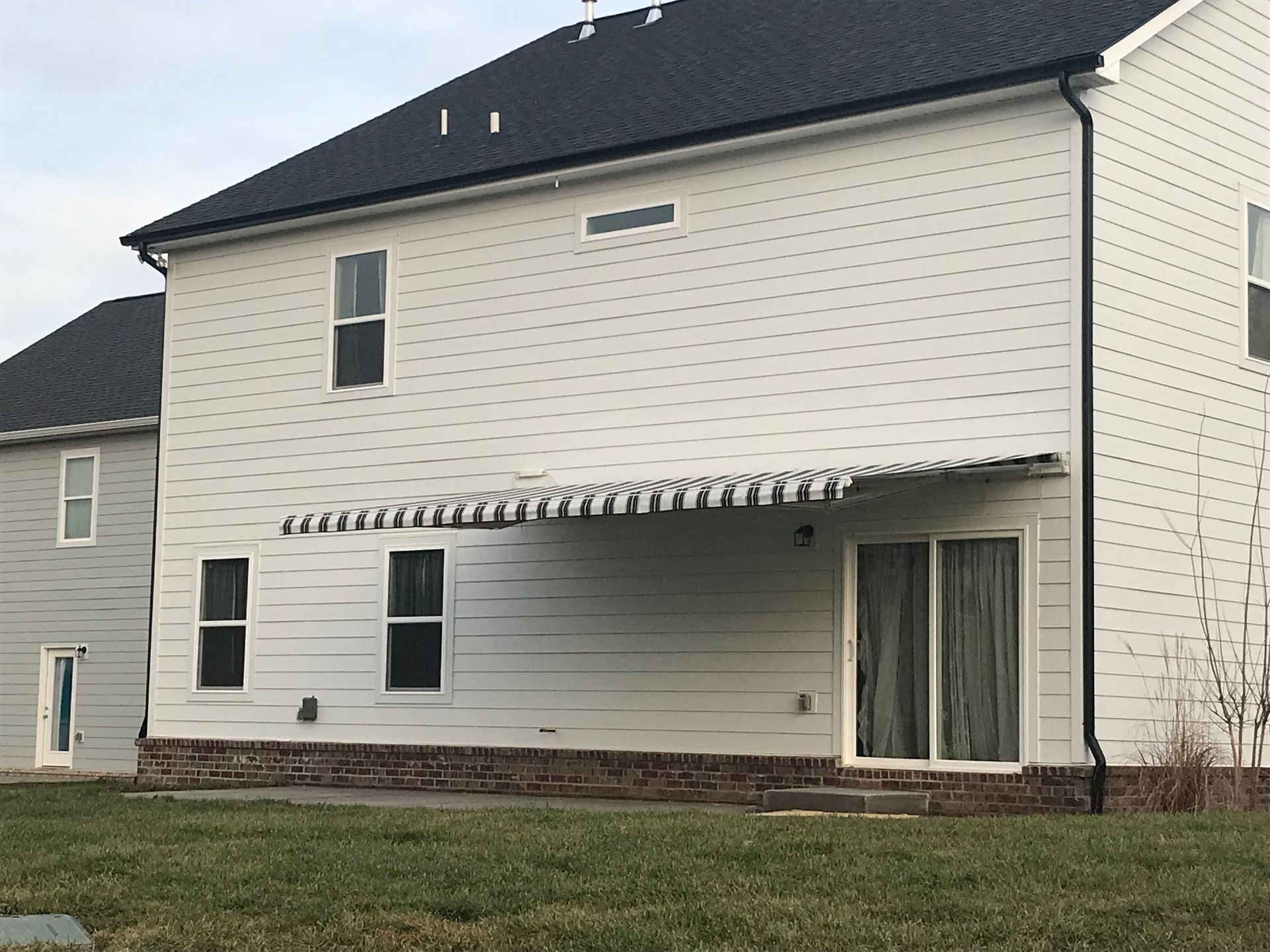 White house with black roof, awning over patio door, two windows, brick base, green grass.