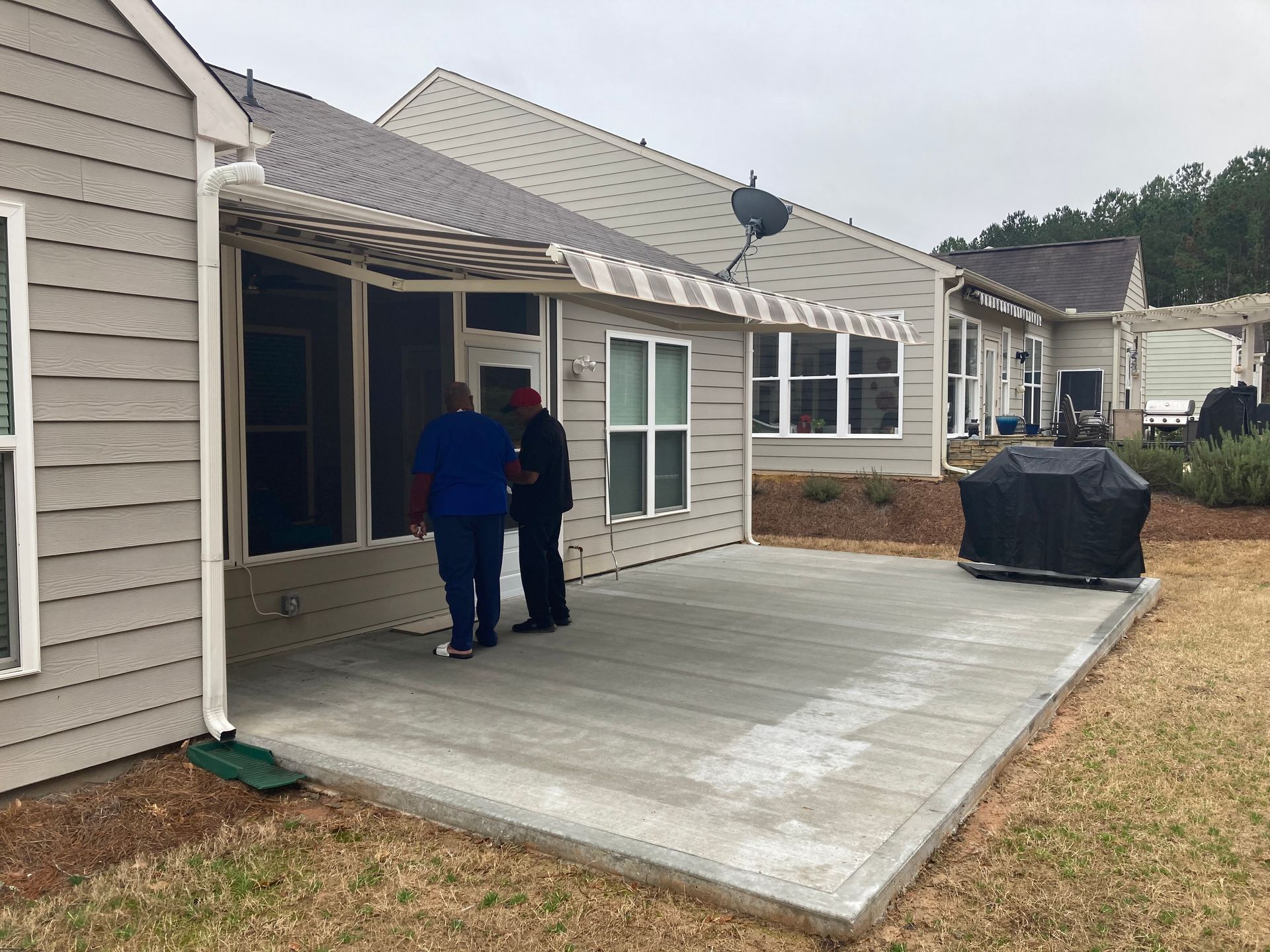 Two people on a gray patio next to a house with an awning, overcast sky.