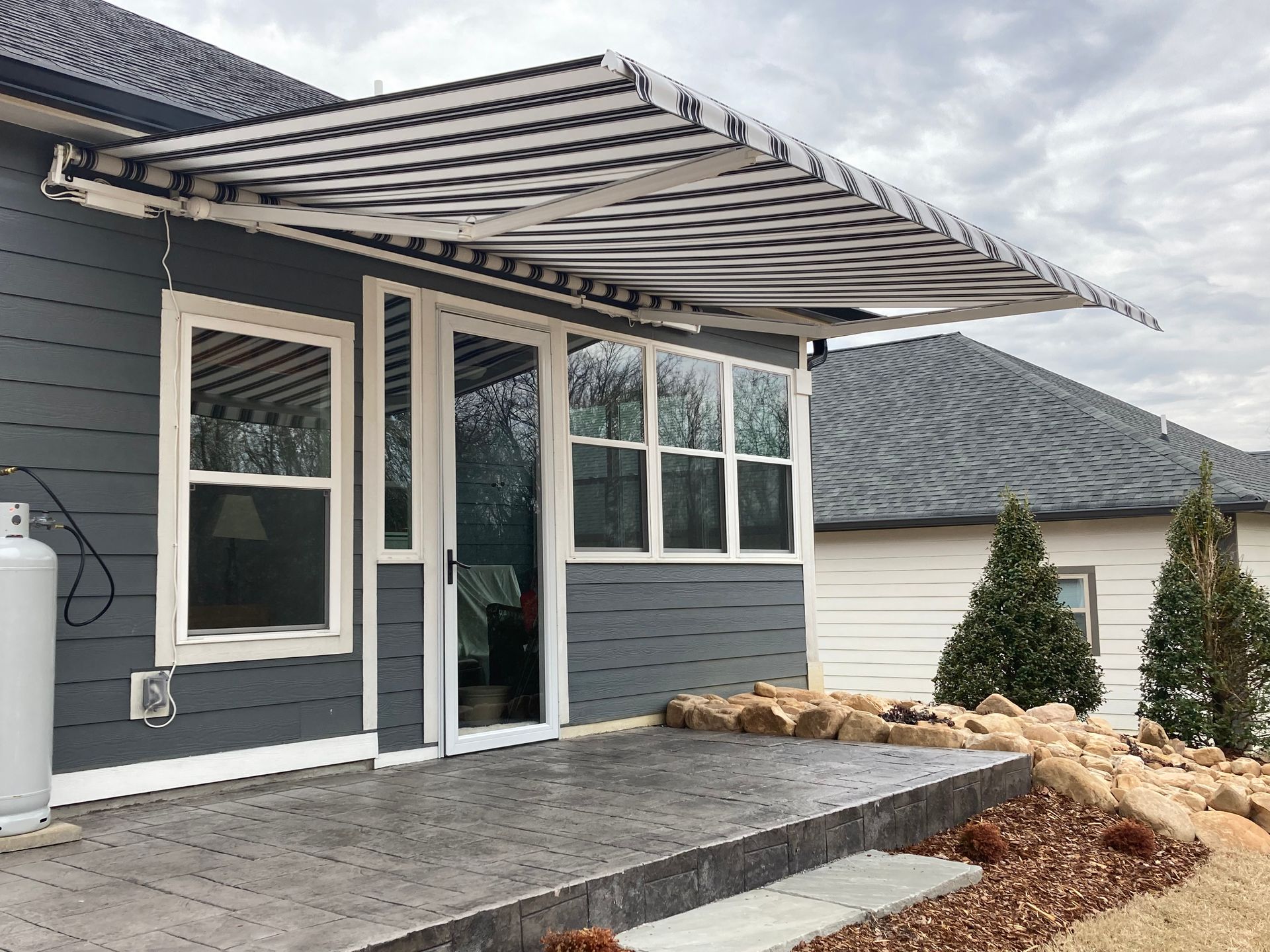 Gray house with striped awning over doors and windows; concrete patio.
