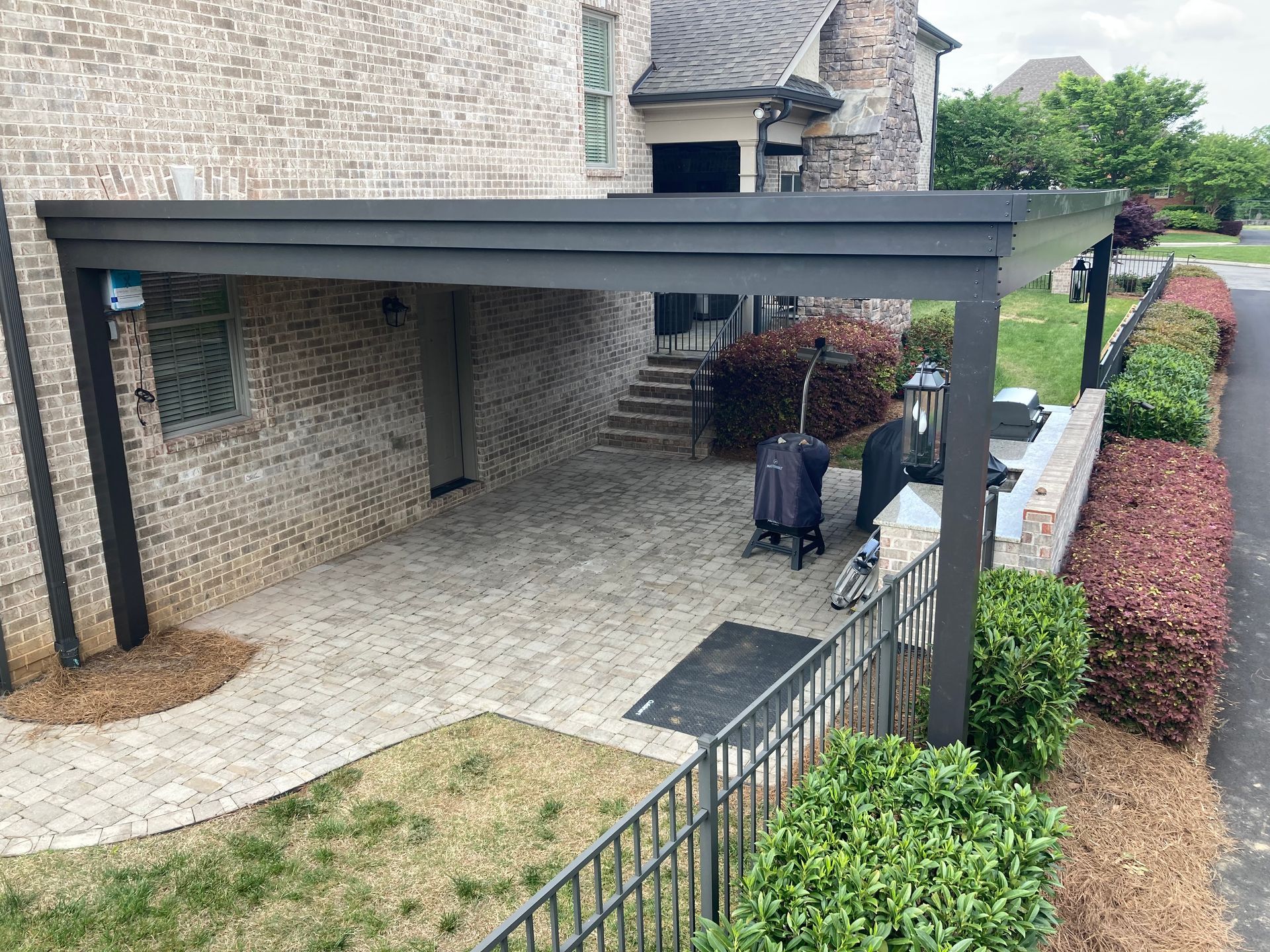 Patio with gray pergola attached to a brick building, paved with pavers, with landscaping.