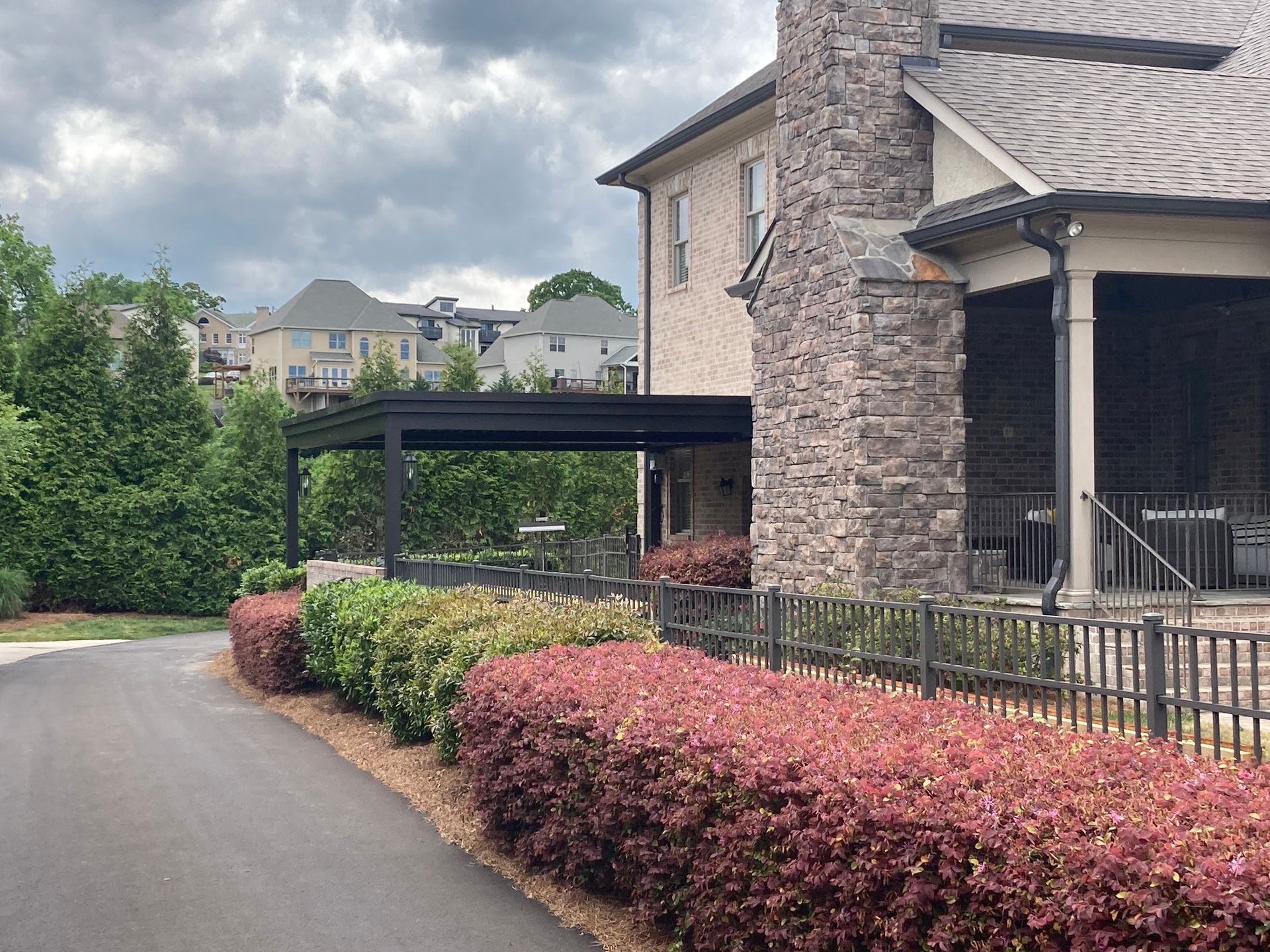 A stone-and-brick house with a black patio cover, red and green bushes, and a paved walkway.