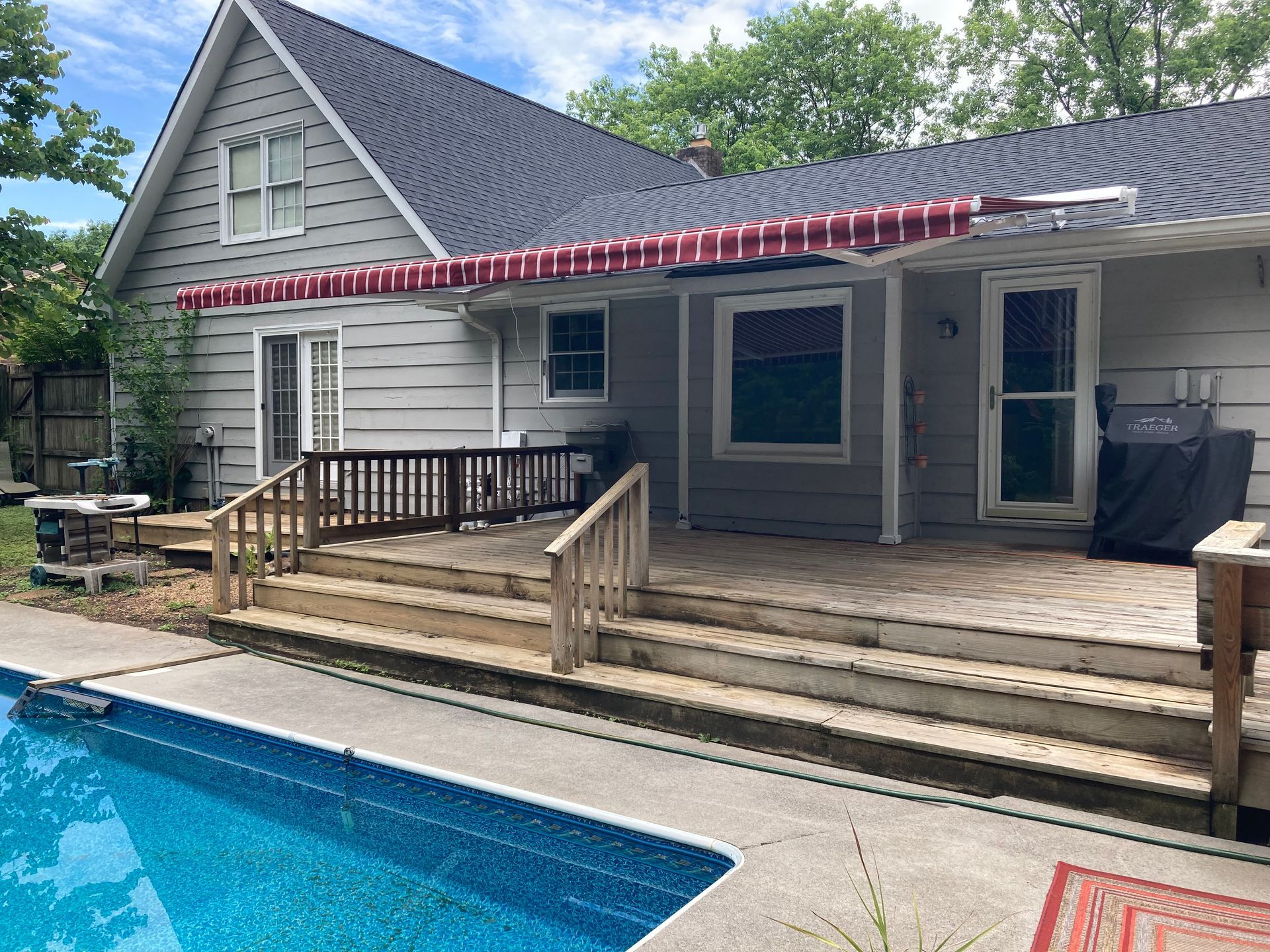 Backyard with a pool, deck, and house. Awnings above windows, with red and white stripes. Sunny day.
